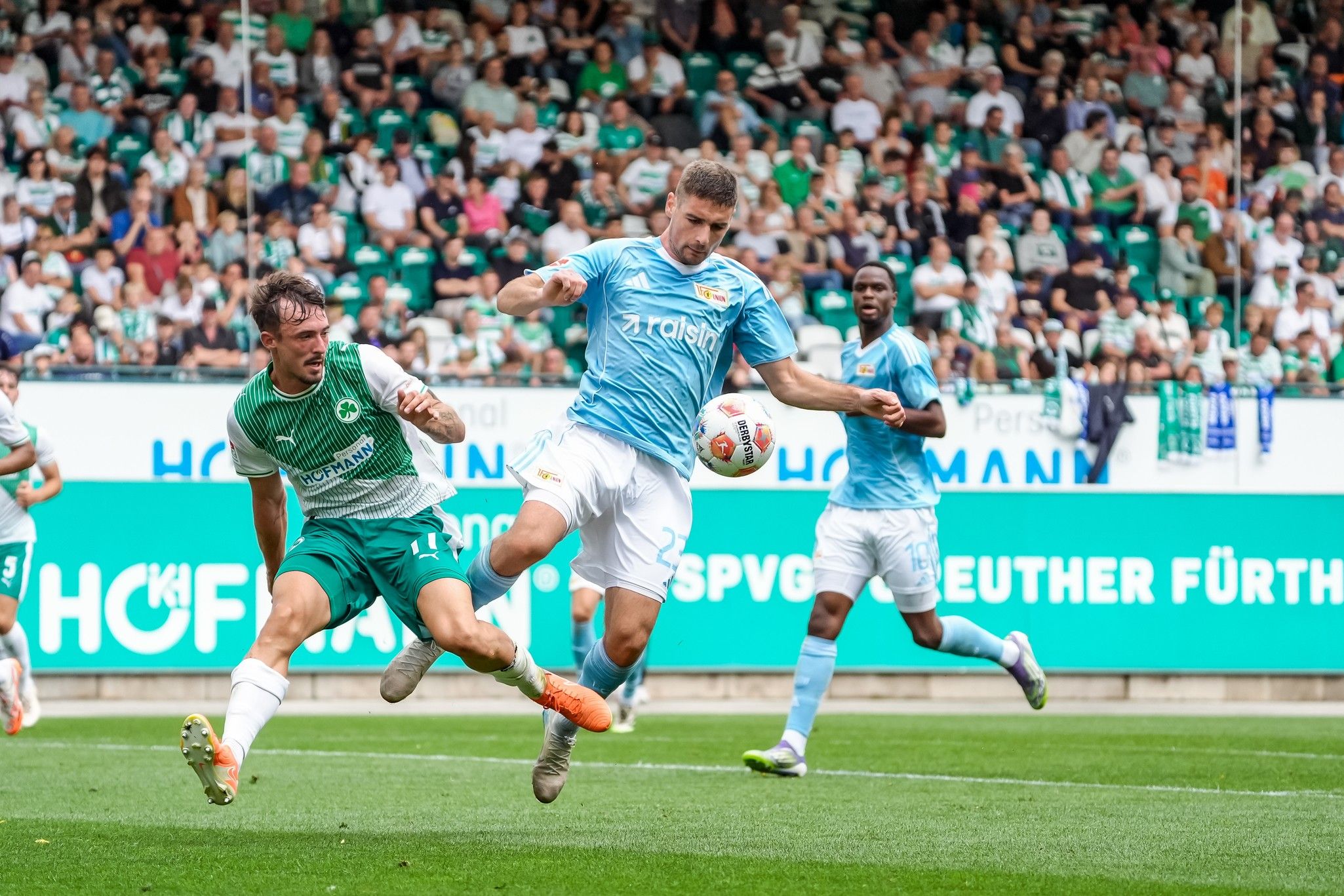 Spielszene im Fußball: Ein Spieler in blauen Trikots schießt den Ball, während ein grüner Spieler versucht, ihn zu blockieren. Zuschauermenge im Hintergrund.