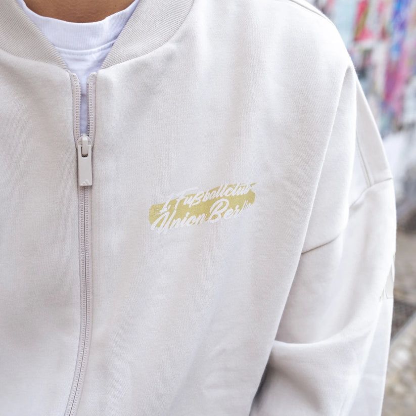 Person trägt eine graue, leicht oversized Jacke mit dem Schriftzug "Union Berlin" in gelber Schrift.