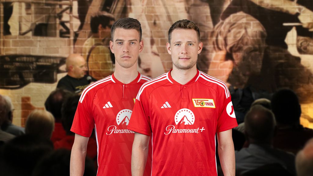 Zwei Fußballspieler in roten Trikots stehen vor einer verschwommenen Hintergrundszene mit Menschenmenge.