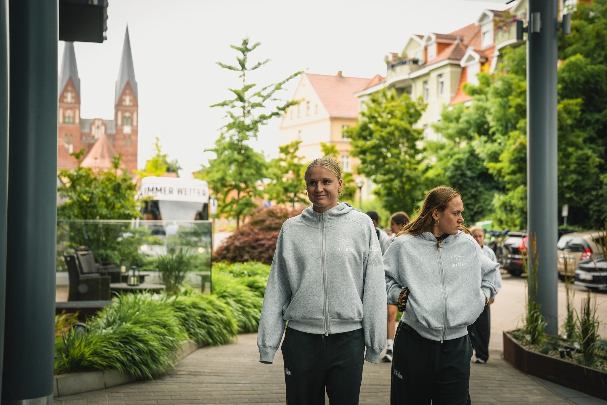 Zwei Personen in grauen Sweatshirts gehen gegenüber einer Häuserreihe mit Bäumen und einer Kirche im Hintergrund.