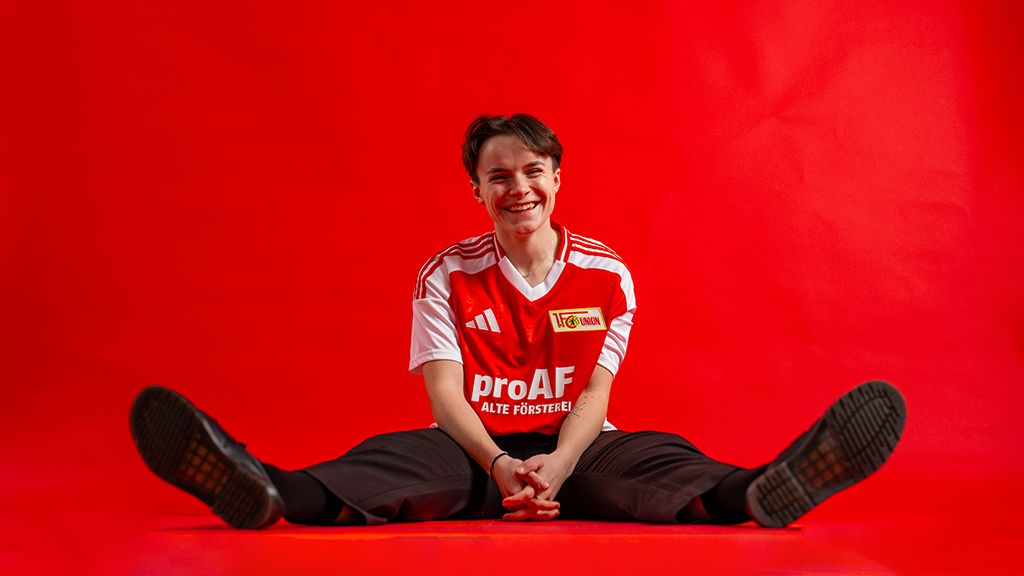 A person in a red and white soccer jersey sits with a smiling face in front of a solid red background.