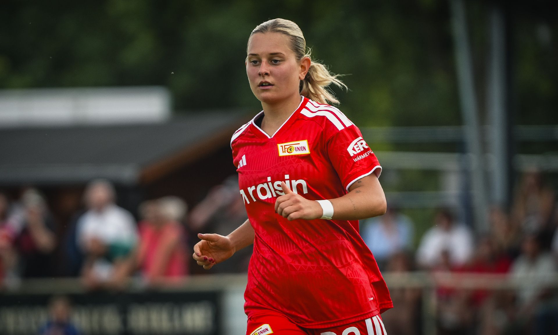 Eine Frau in einem roten Fußballtrikot joggt auf dem Spielfeld, im Hintergrund sind Zuschauer zu sehen.