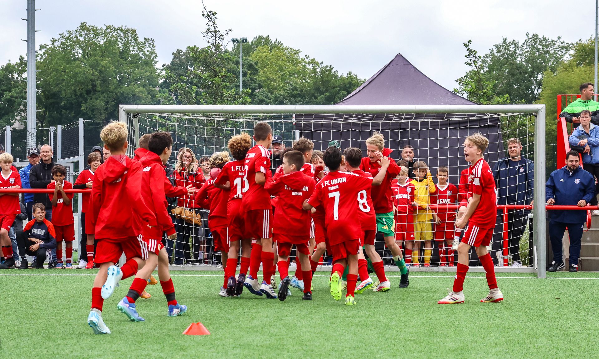Gruppierung von jungen Fußballspielern in roten Trikots, die nach einem Torjubel auf einem Spielfeld feiern. Zuschauer im Hintergrund.