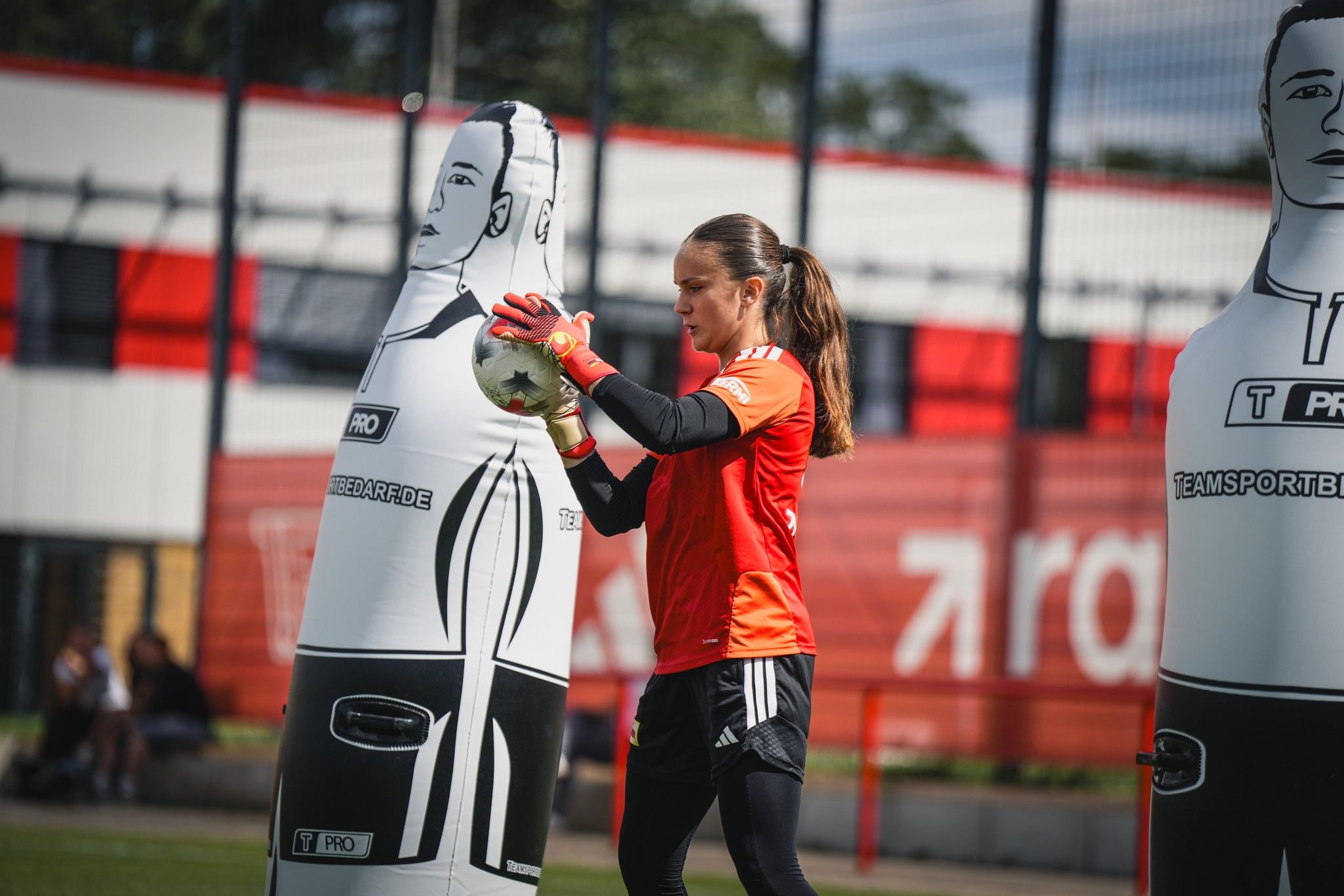 Eine Torhüterin in orangefarbener Ausrüstung trainiert mit einem Trainingsdummy auf einem Fußballplatz.