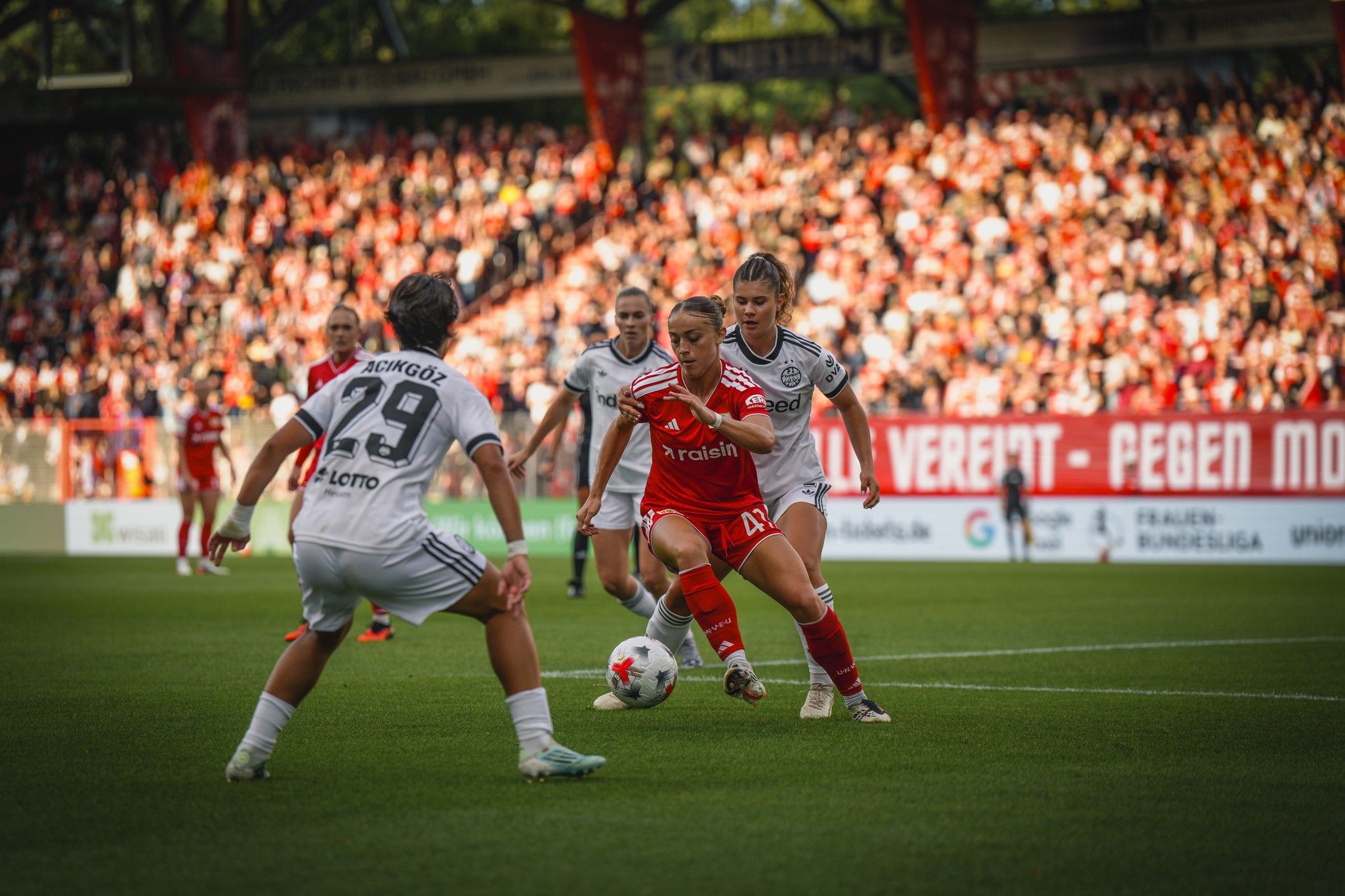 Fußballspiel mit Spielerinnen in roten und weißen Trikots, die um den Ball kämpfen, vor jubelndem Publikum.