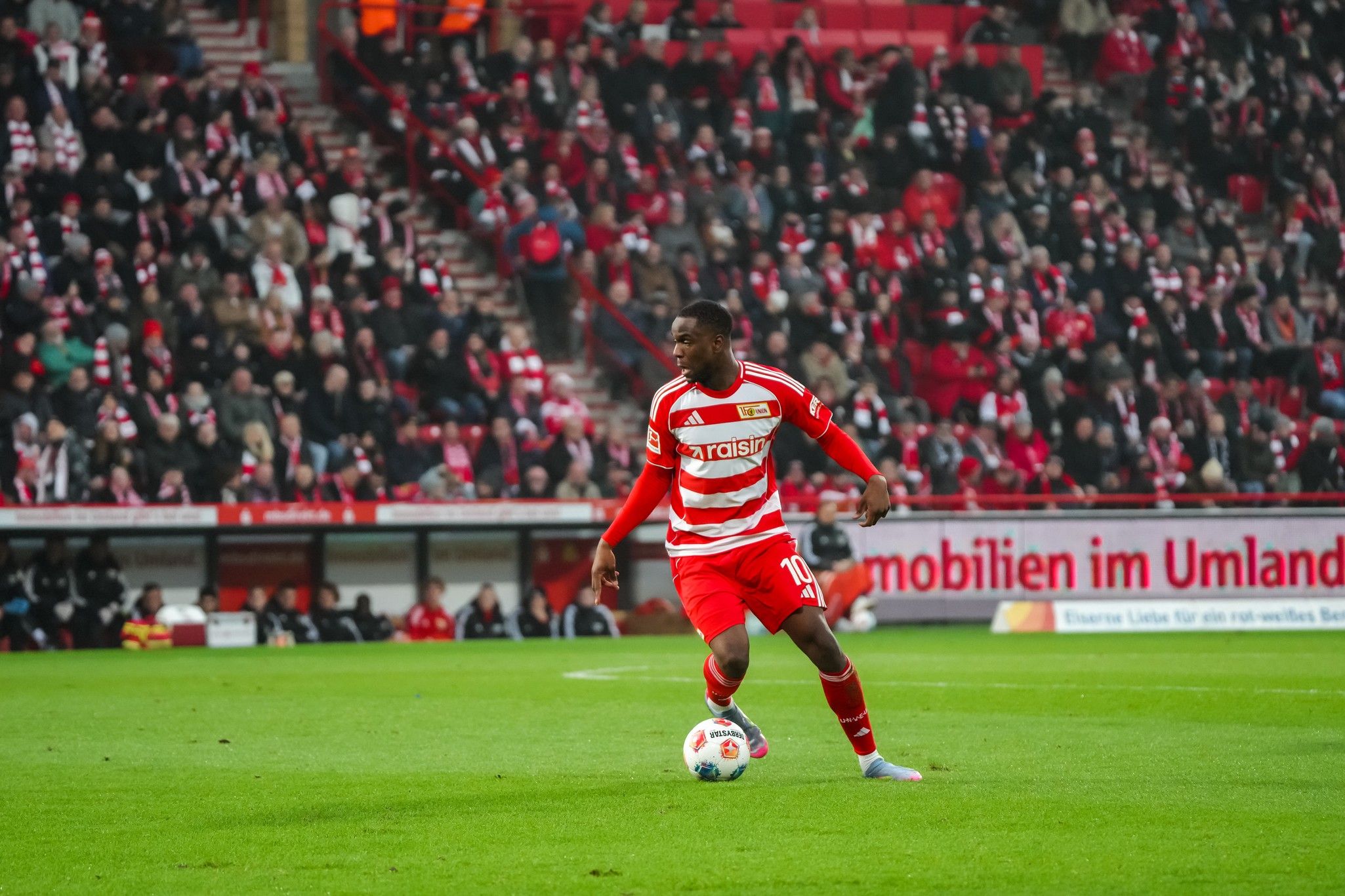 Ein Spieler in rotem Trikot mit Streifen dribbelt den Ball auf dem Fußballfeld, im Hintergrund eine jubelnde Menge.