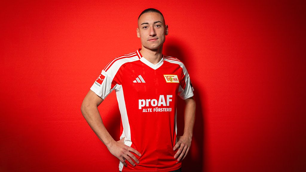 Ein junger Mann in einem roten Fußballtrikot mit einem weißen Logo steht vor einem roten Hintergrund.