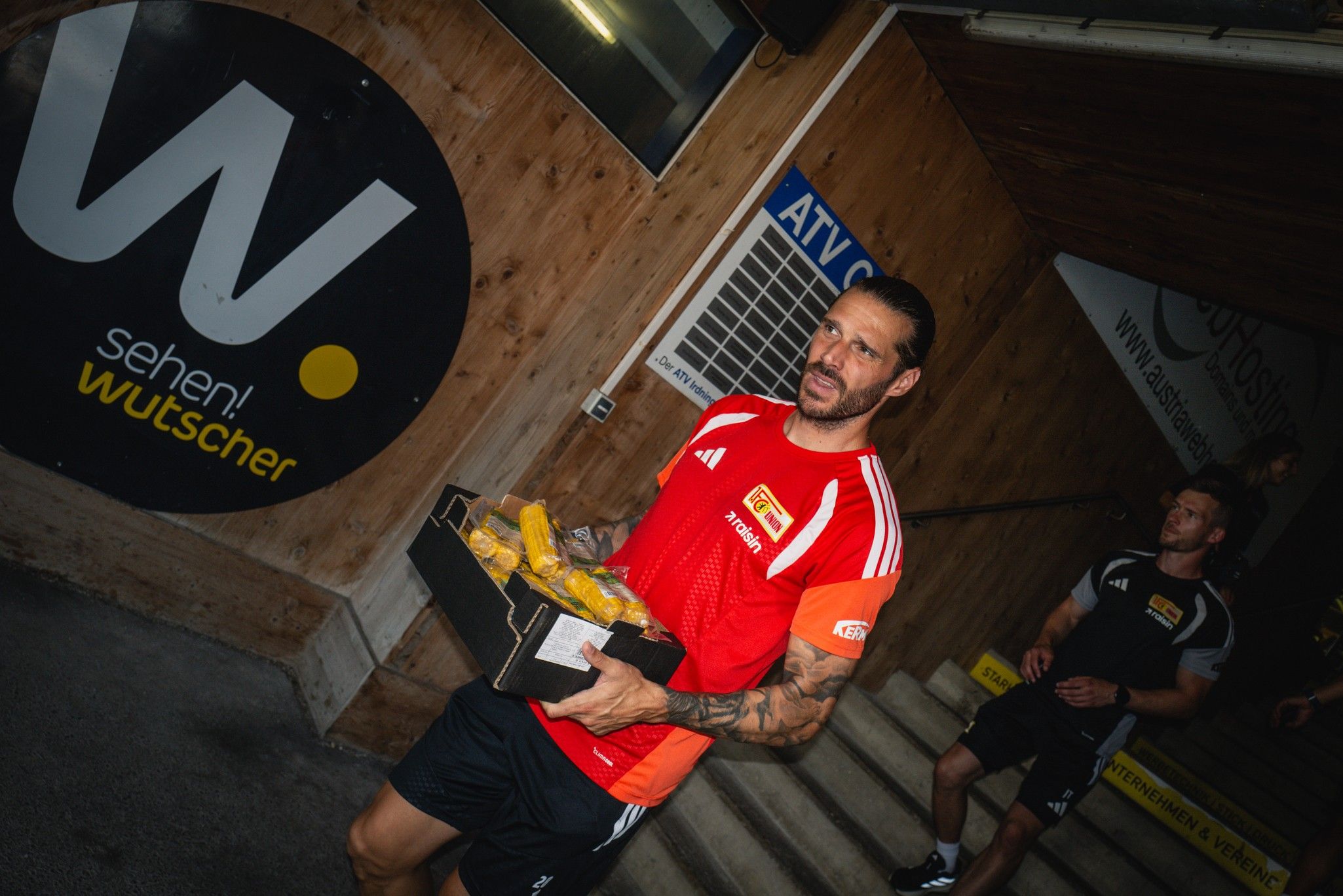 Ein Fußballspieler trägt einen Kasten mit Getränken, während er eine Treppe hinuntergeht. Im Hintergrund sind weitere Spieler sichtbar.