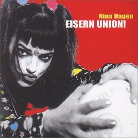 Künstlerin Nina Hagen mit auffälligem Make-up und buntem Outfit hält einen Fußball vor rotem Hintergrund.