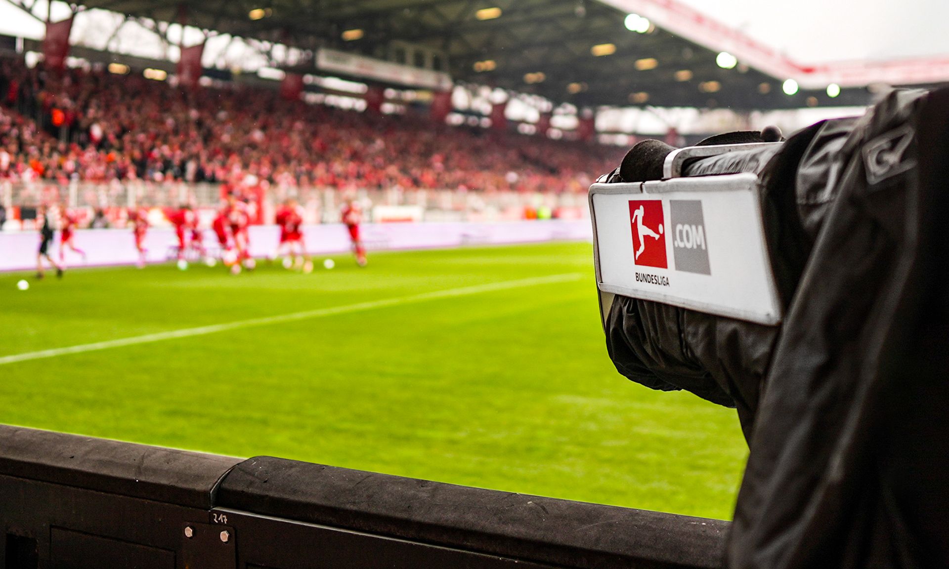 Kamera eines Kameramanns im Vordergrund eines Fußballspiels, im Hintergrund sind Spieler und Zuschauer auf dem Stadiongelände zu sehen.