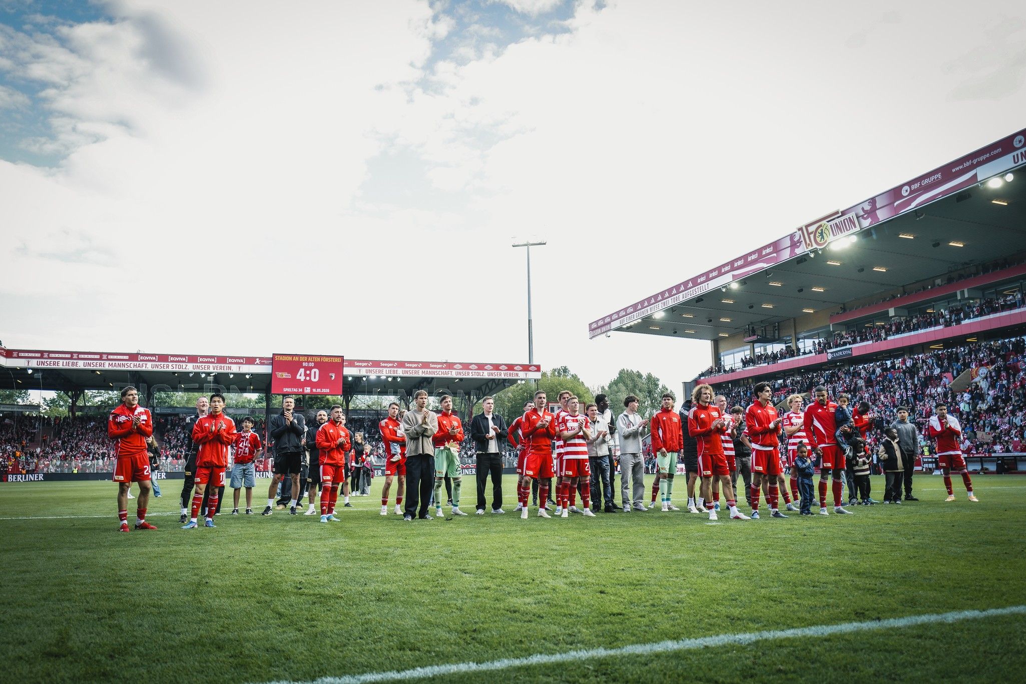 Feiernde Fußballmannschaft in roten Trikots auf dem Spielfeld, Zuschauer im Hintergrund. Stadionatmosphäre mit jubelnden Fans.