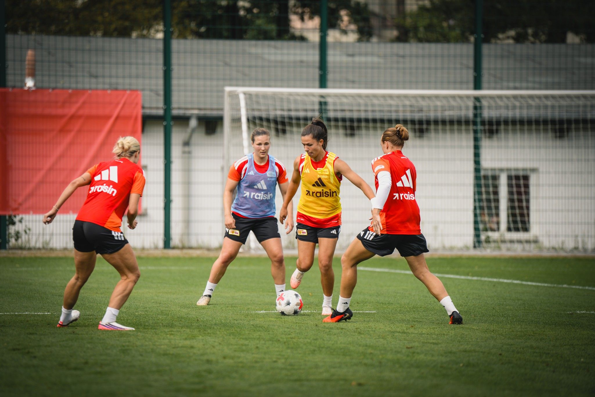Vier weibliche Fußballspielerinnen trainieren auf einem Rasenfeld, zwei tragen rote Trikots, eine ein gelbes Trikot.