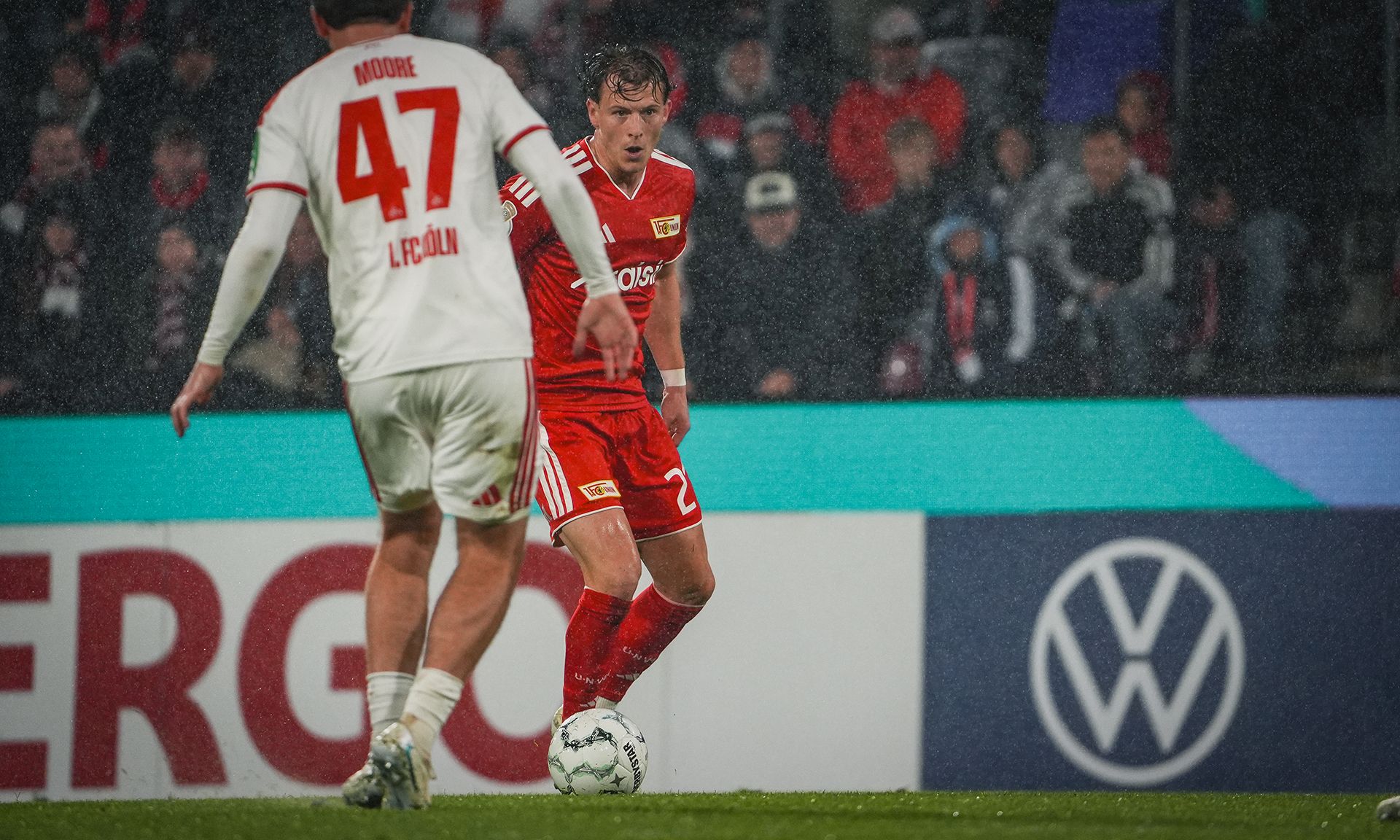 Zwei Fußballspieler auf dem Platz während eines Spiels, einer in rot, der andere in weiß, bei Regen.