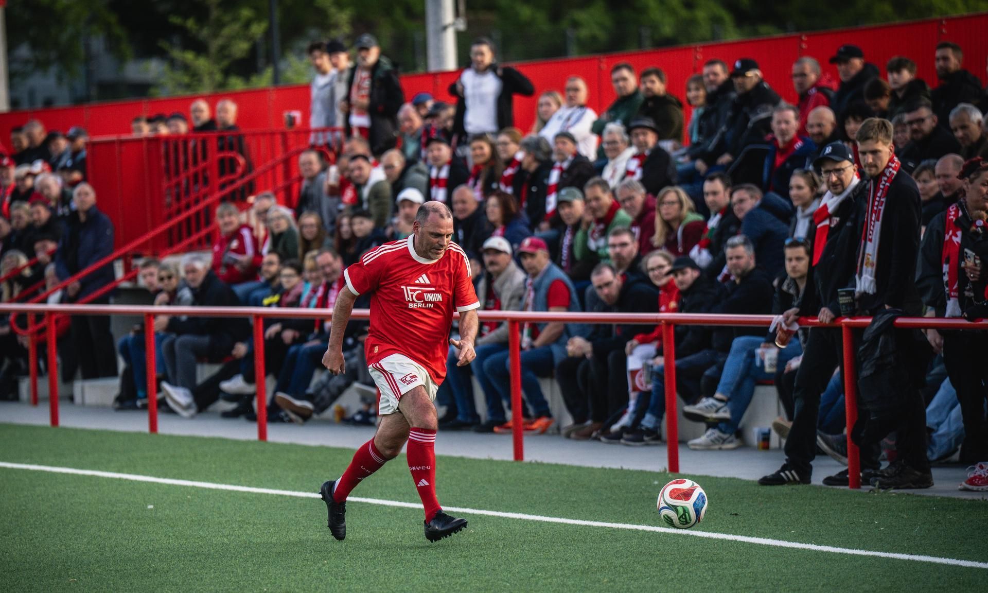 Ein Fußballspieler in rotem Trikot führt einen Einwurf aus, während Zuschauer auf den Tribünen aufmerksam zuschauen.