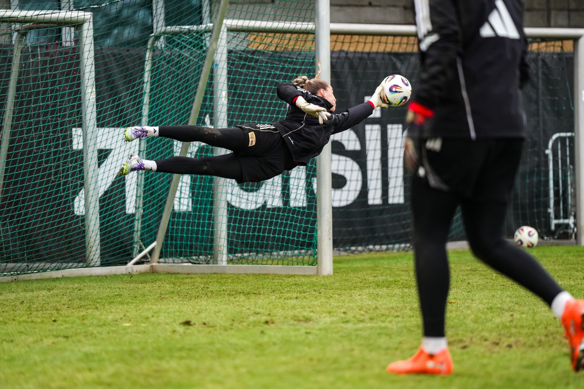 Torhüterin springt nach rechts, um einen Fußball während des Trainings zu halten. Im Hintergrund ist ein weiteres Spieler sichtbar.