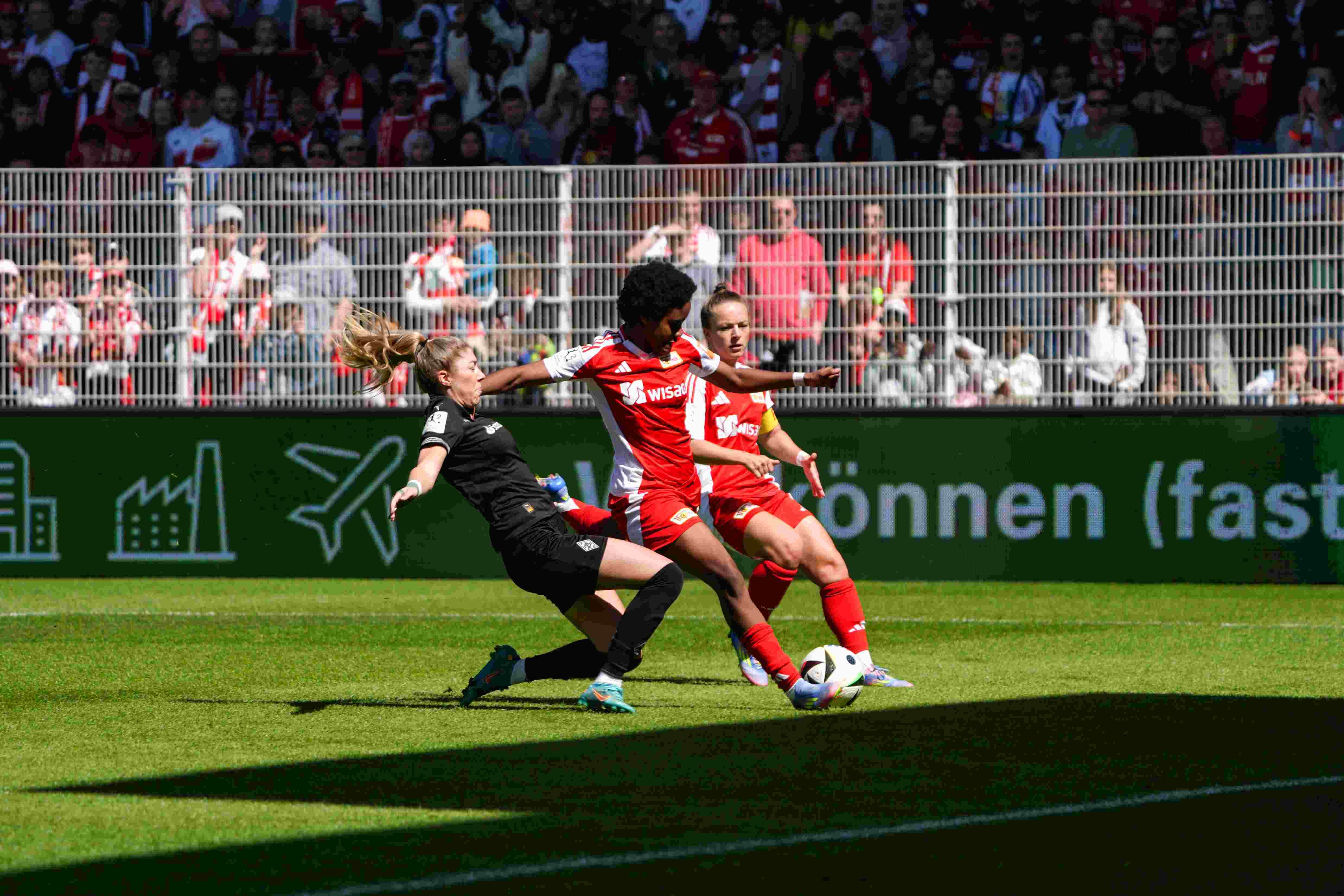 Zwei Fußballspielerinnen in roten Trikots kämpfen um den Ball, während eine Torhüterin in Schwarz am Boden liegt.