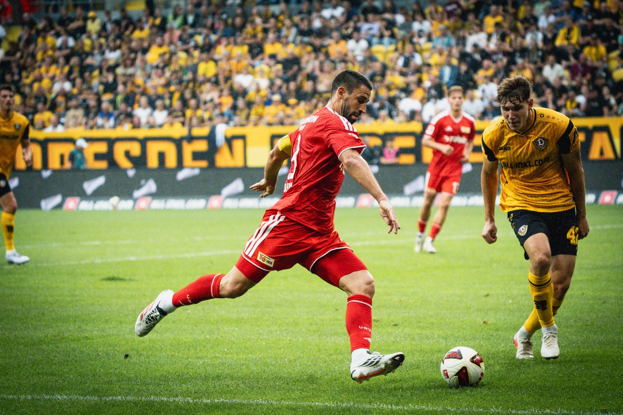 Ein Fußballspieler in roter Uniform tritt den Ball, während ein gegnerischer Spieler in gelber Uniform ihn verfolgt. Zuschauer im Hintergrund.