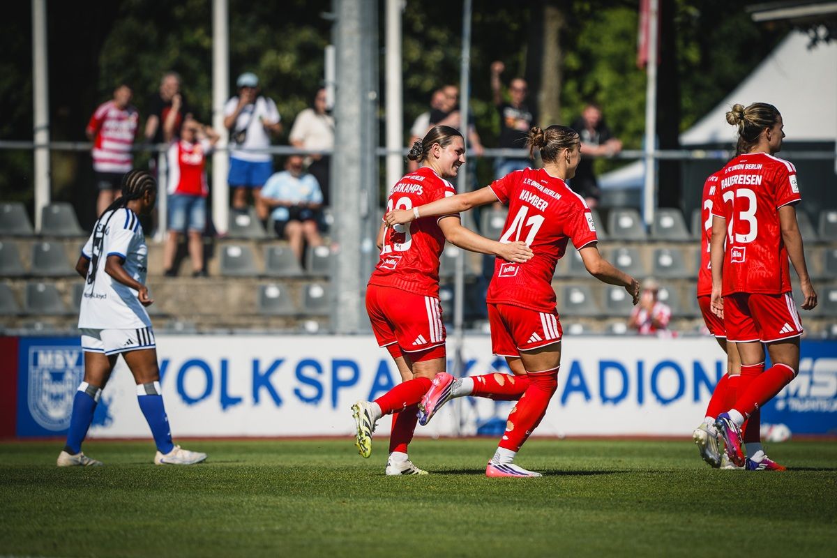 Frauenfußballspiel mit Spielerinnen in roten Trikots, die jubeln, während eine andere Spielerin in Blau zuschaut.