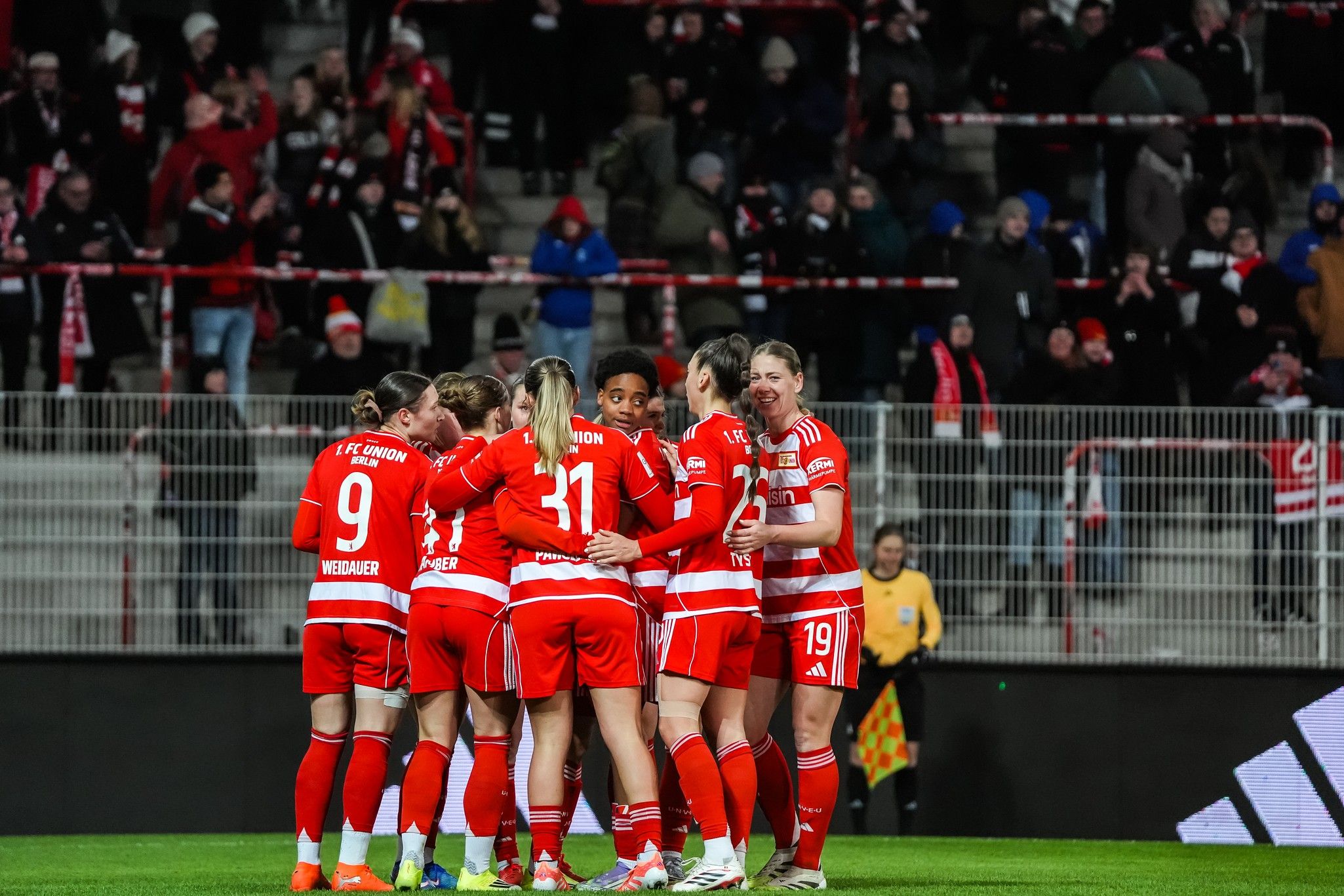 Spielerinnen eines Fußballteams in roten Trikots feiern gemeinsam auf dem Feld vor jubelnden Fans.