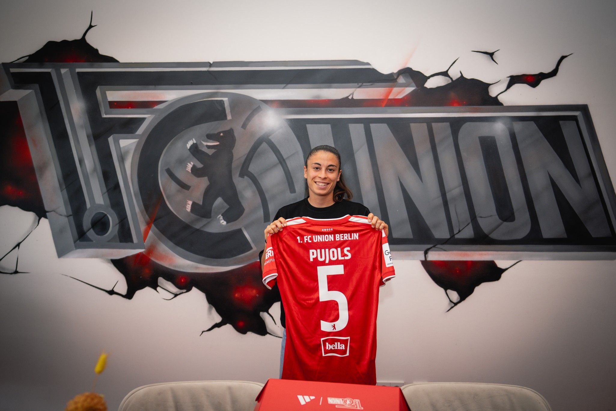 Frau hält ein Trikot mit der Nummer 5 von FC Union Berlin vor einem großen Vereinslogo.