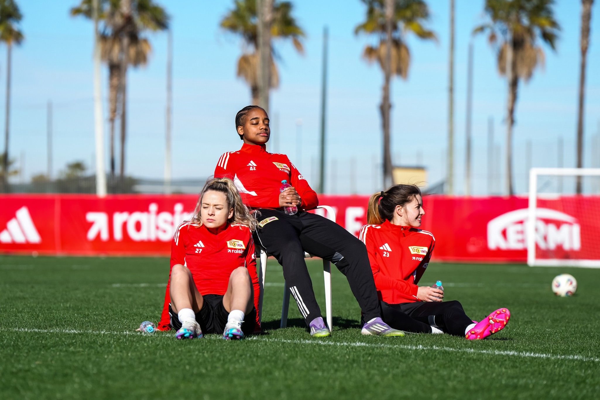 Drei Fußballspielerinnen in roten Trainingsanzügen sitzen auf dem Rasen, umgeben von Palmen und einem Fußballtor im Hintergrund.