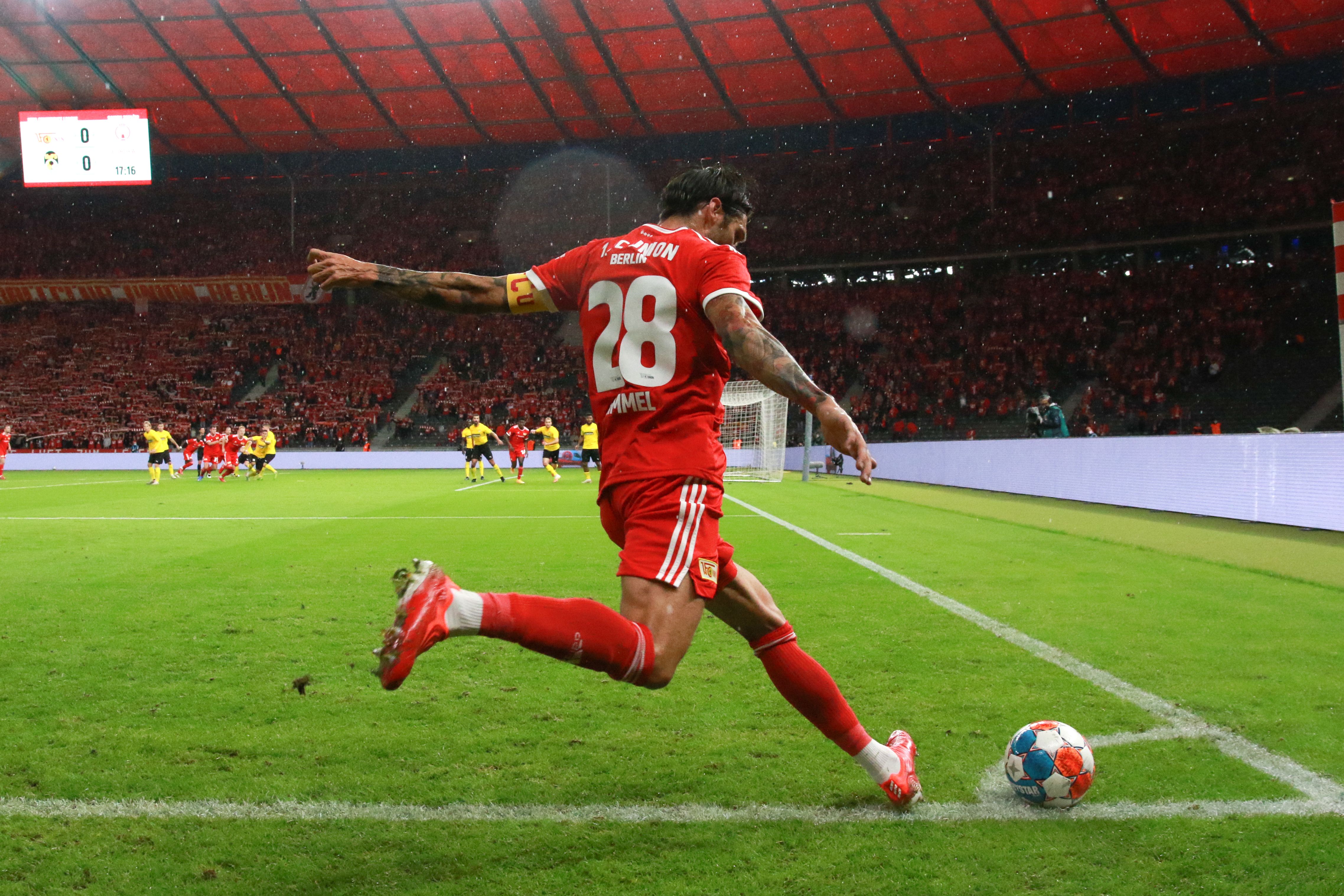 A soccer player in red gear takes a corner kick in a stadium with cheering fans in the background.