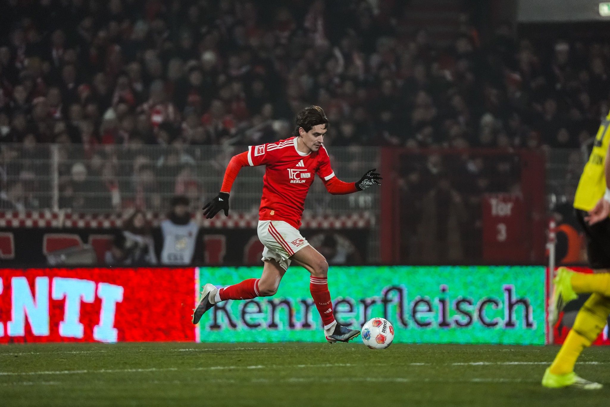 Ein Fußballspieler in roter Kleidung dribbelt den Ball auf dem Spielfeld, umgeben von jubelnden Fans im Hintergrund.