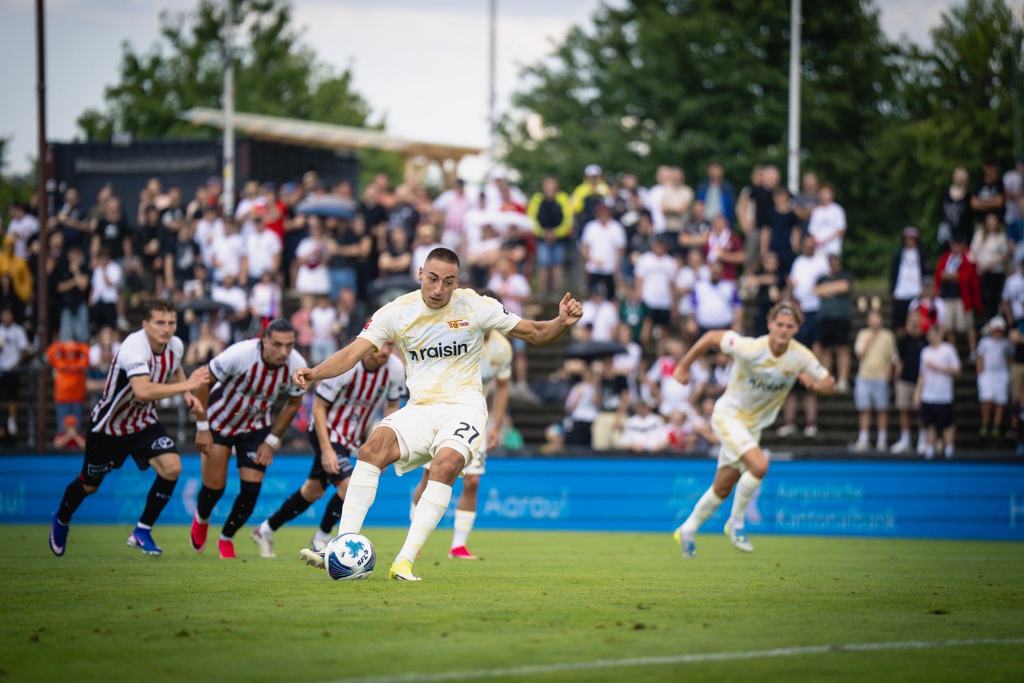 Ein Fußballspieler schießt einen Strafstoß, während Zuschauer im Hintergrund zuschauen.