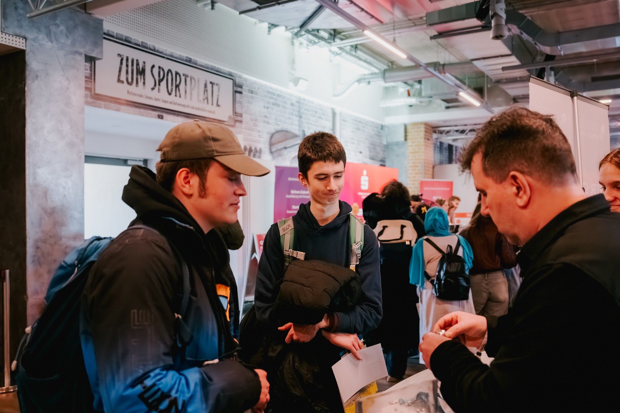 Zwei Personen in winterlichen Jacken stehen an einem Messestand, während ein Gesprächspartner ihnen Informationen gibt.