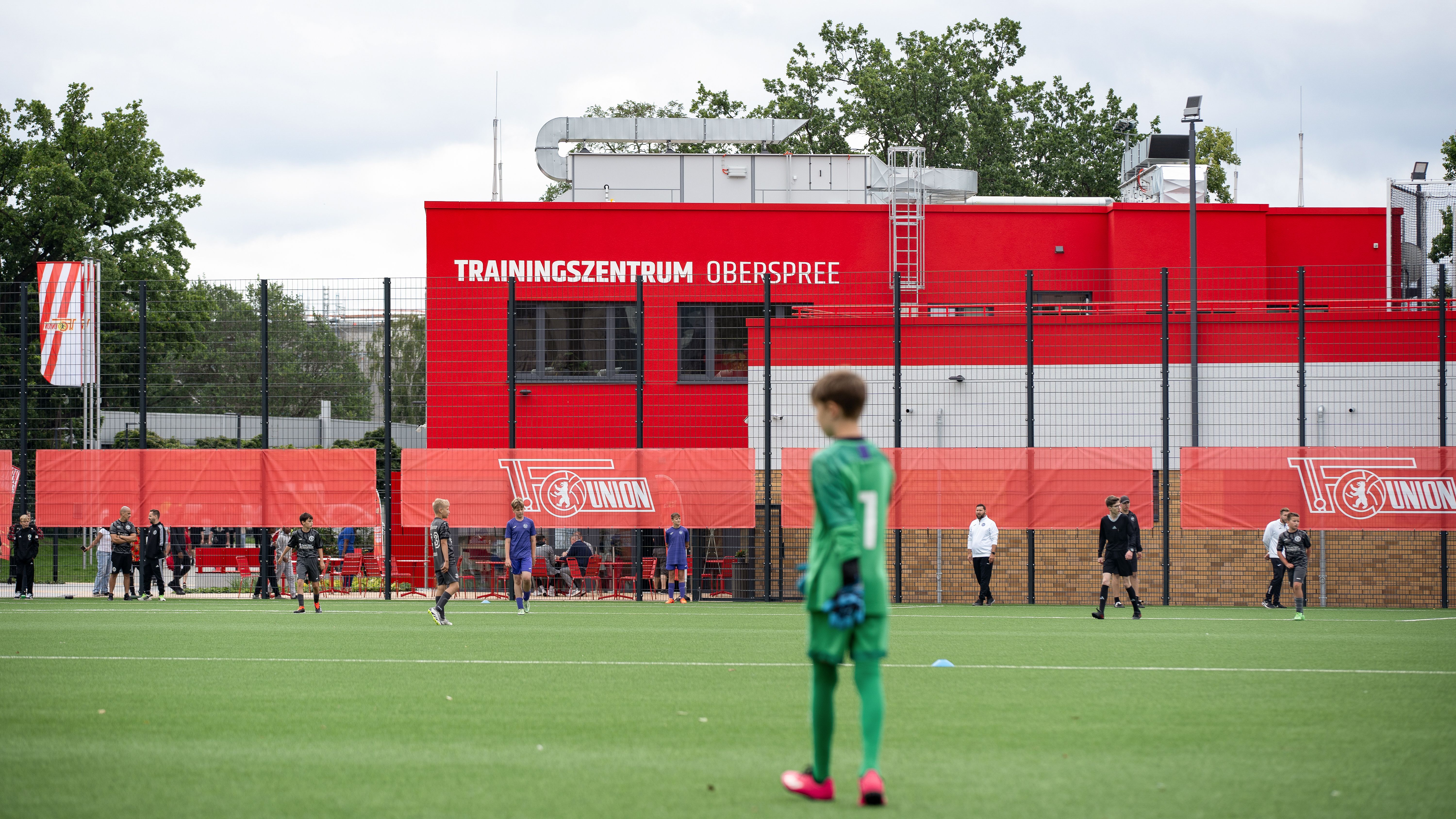 Ein Fußballplatz mit Spielern im Training und einem roten Gebäude im Hintergrund, beschriftet mit "TRAININGZENTRUM OEBERSPOREE".