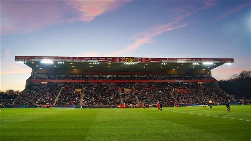 Blick auf ein voll besetztes Fußballstadion bei Sonnenuntergang, mit einem Grasfeld und Spielern im Vordergrund.