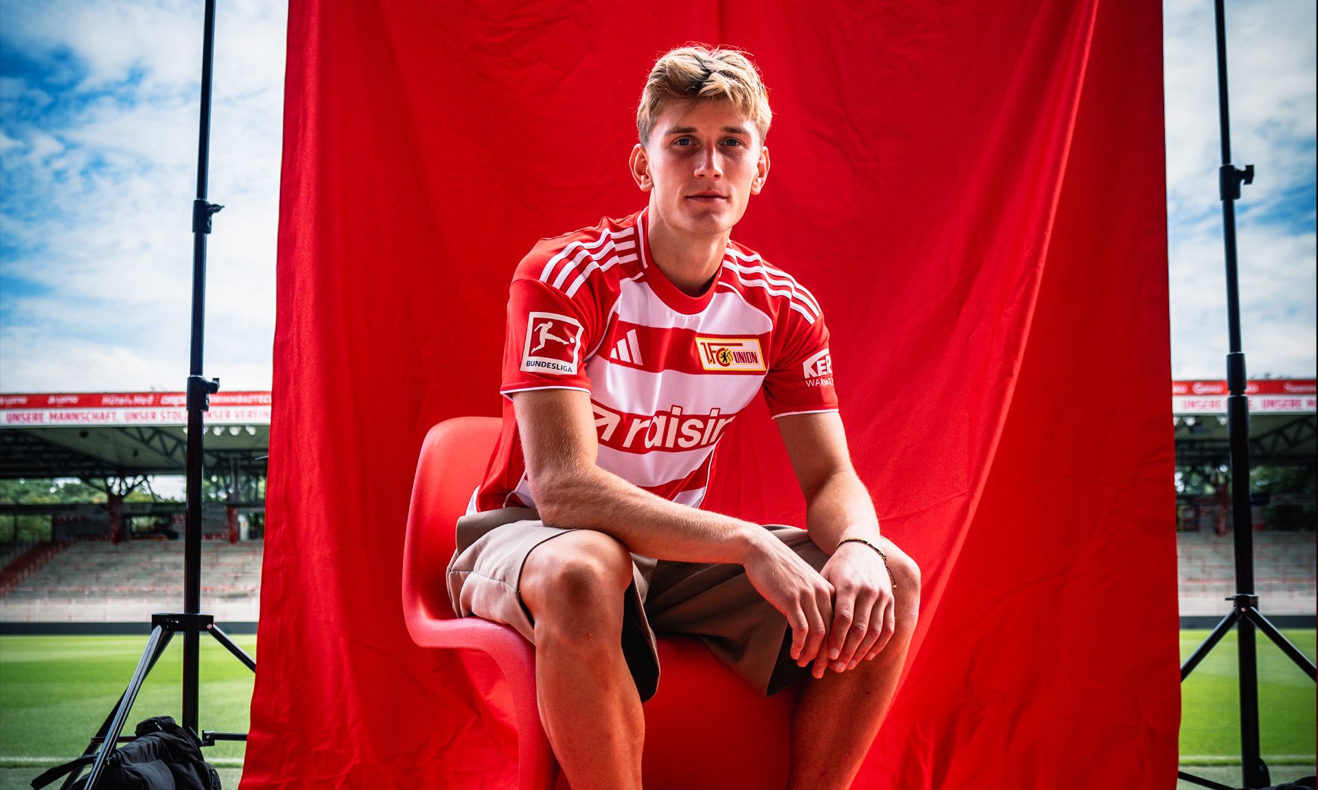 Junger Fußballspieler in rotem Trikot sitzt auf einem Stuhl vor einem roten Hintergrund in einem Stadion.