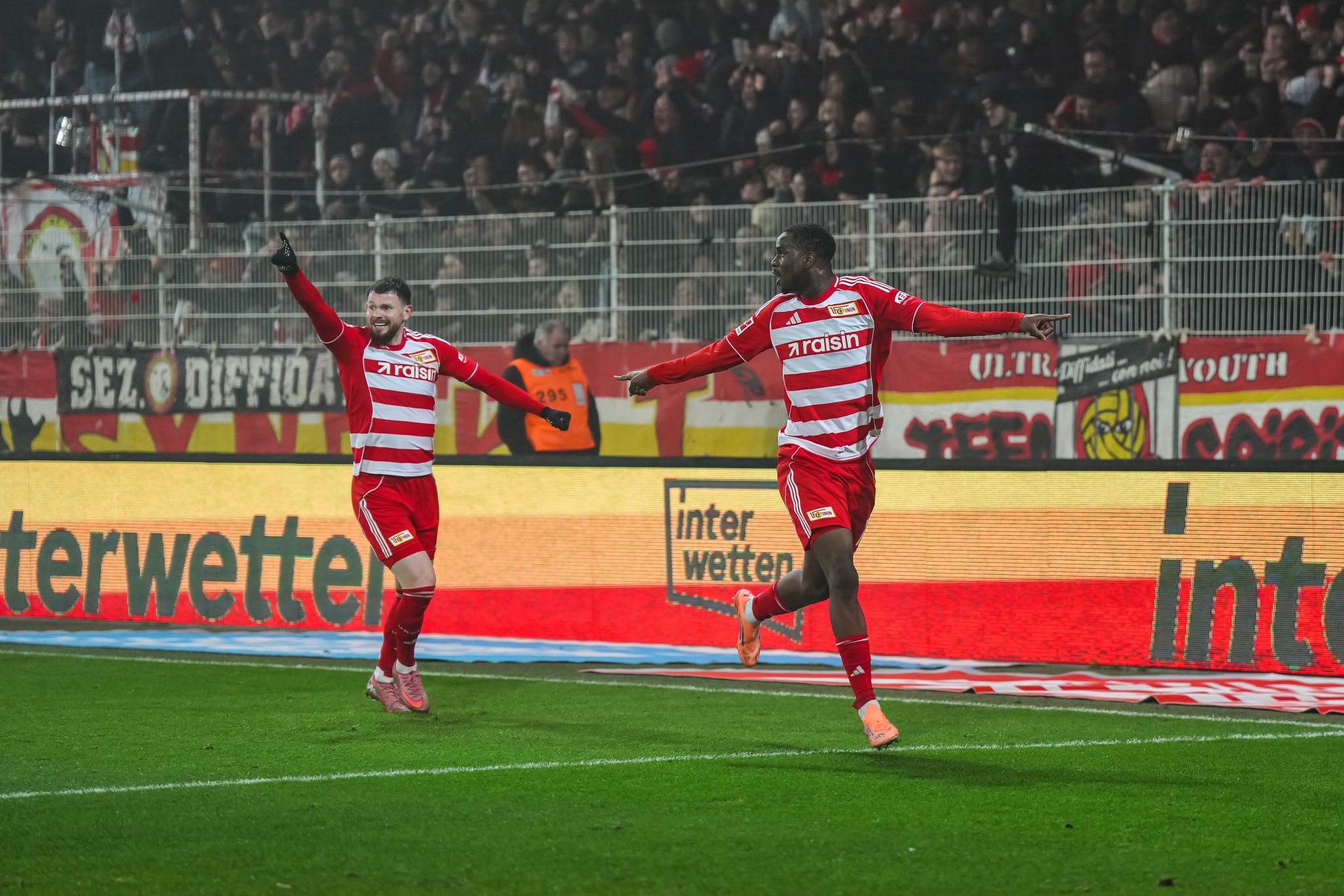 Zwei Fußballspieler in rot-weißen Trikots jubeln nach einem Tor vor jubelnden Fans im Stadion.