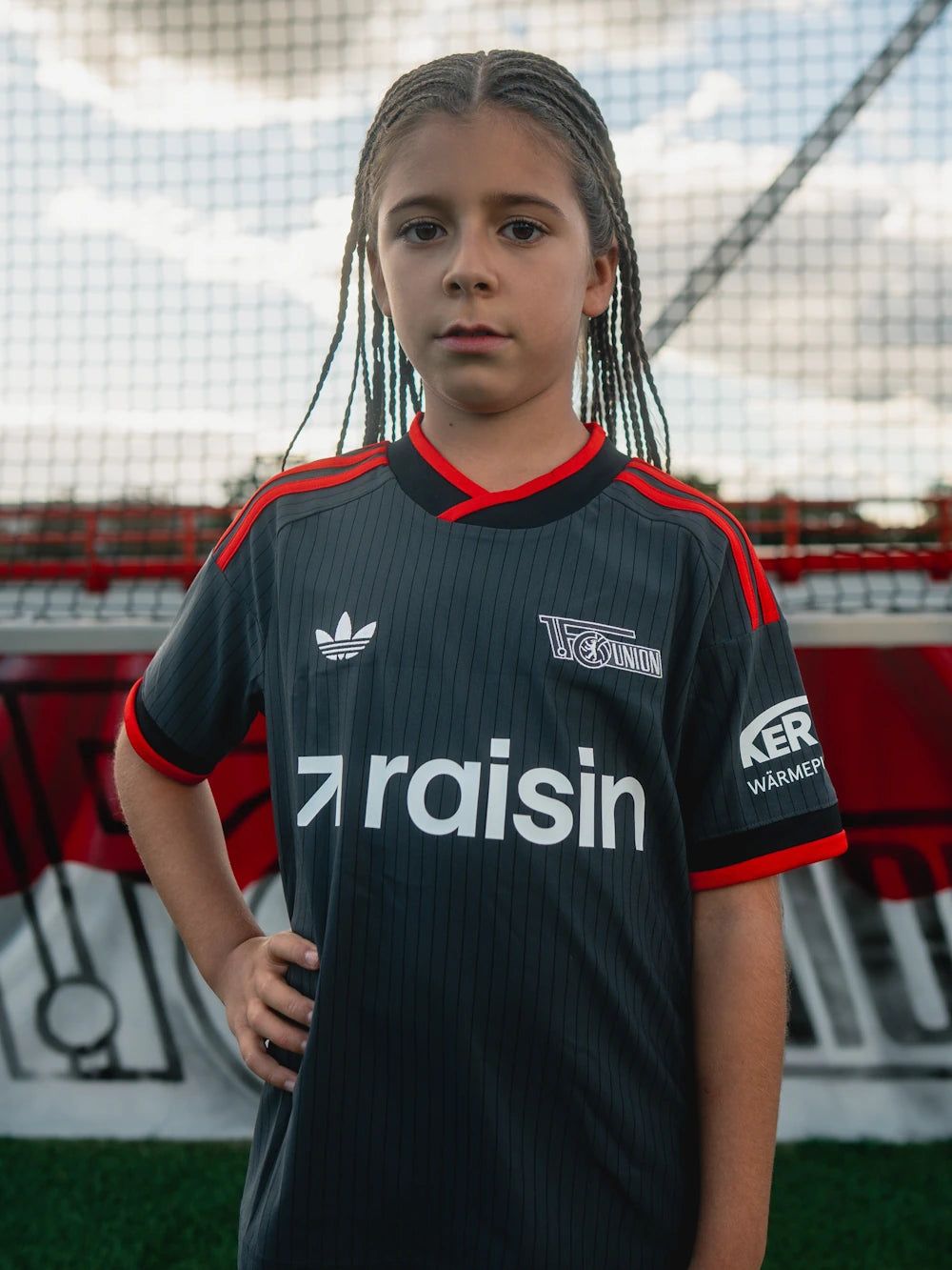 Ein Mädchen mit langen Zöpfen trägt ein dunkelgraues Fußballtrikot mit roten Akzenten und steht auf einem Fußballfeld.