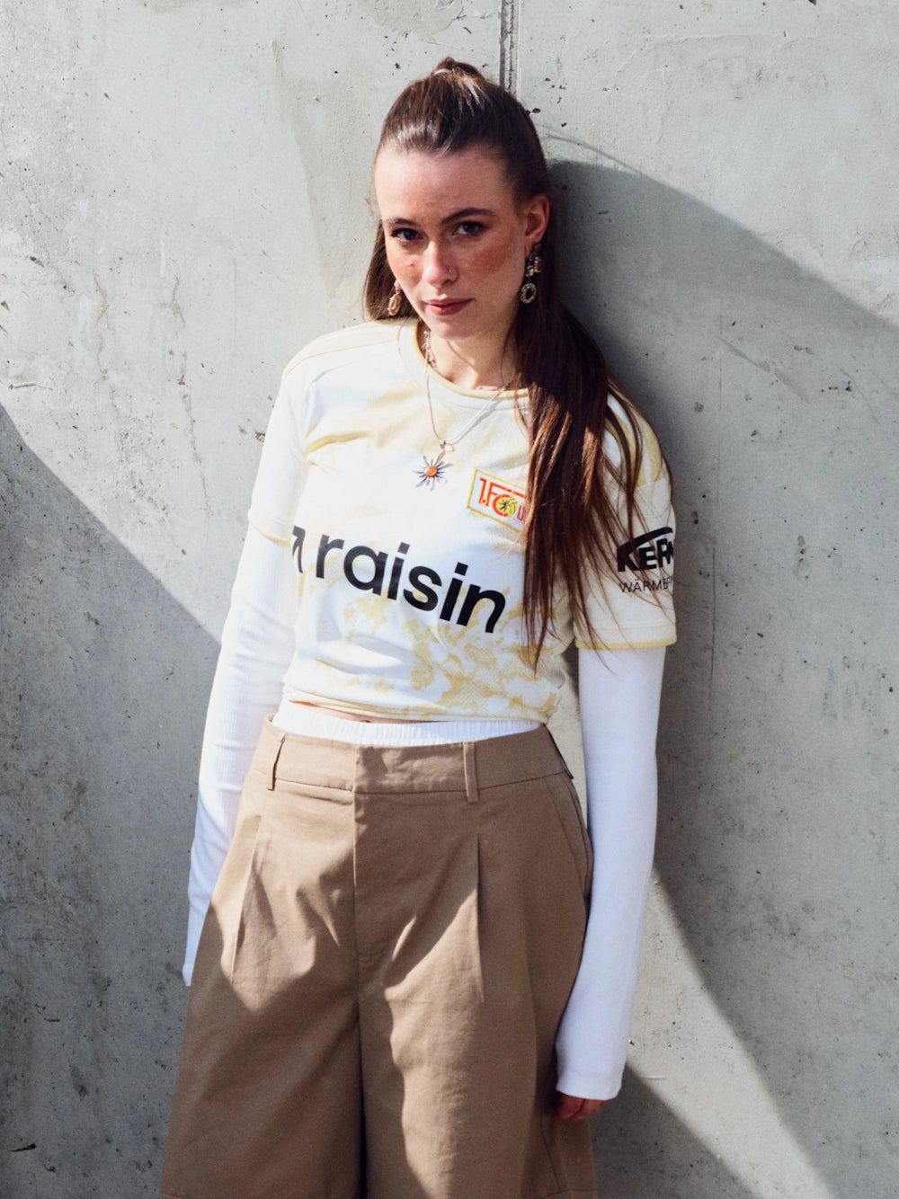 Eine Person in einem Fußballtrikot mit langen Ärmeln und weiten Hosen steht vor einer grauen Wand.