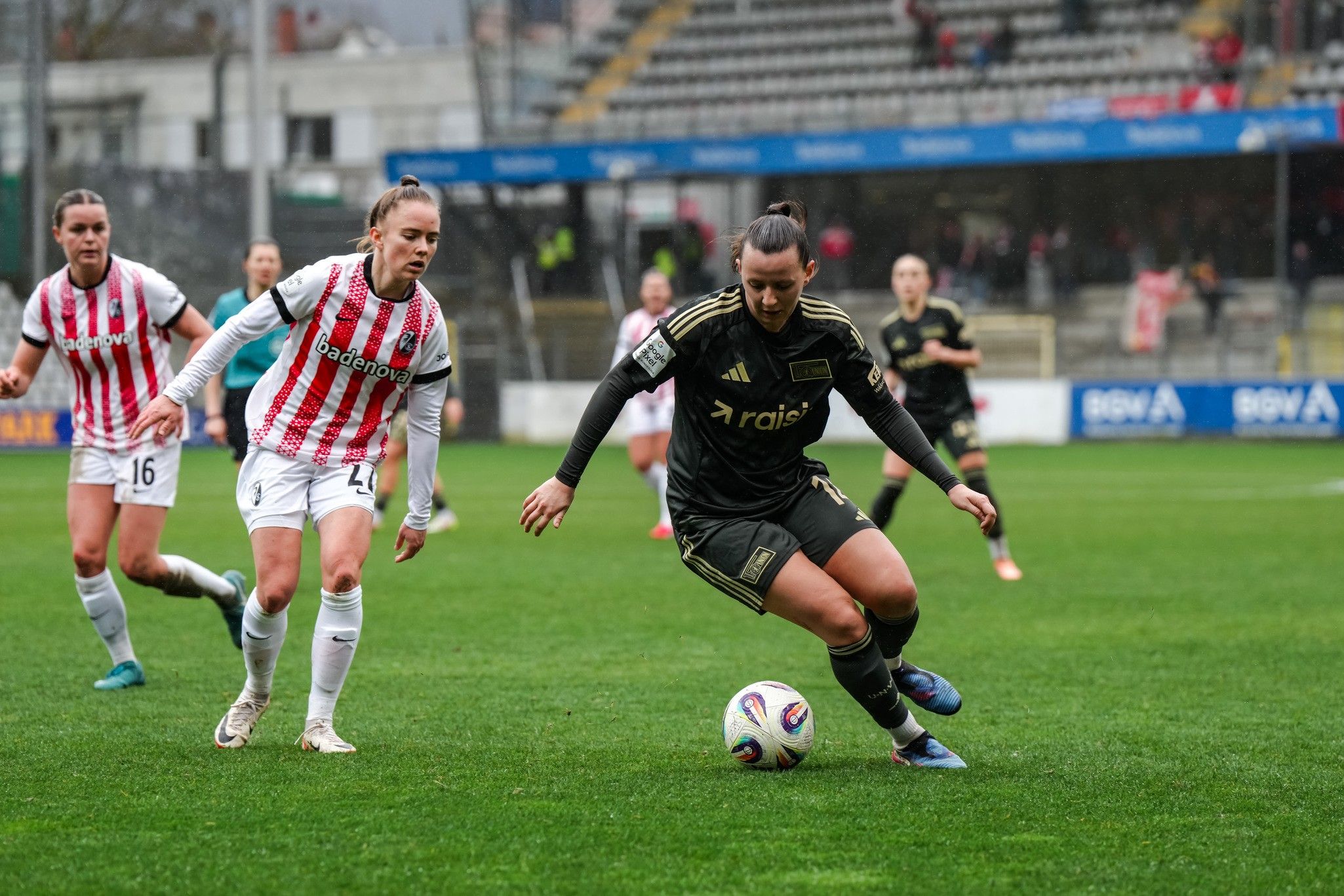 Zwei Fußballspielerinnen im Wettkampf auf dem Spielfeld; eine Spielerin in schwarzer, die andere in gestreifter Uniform.