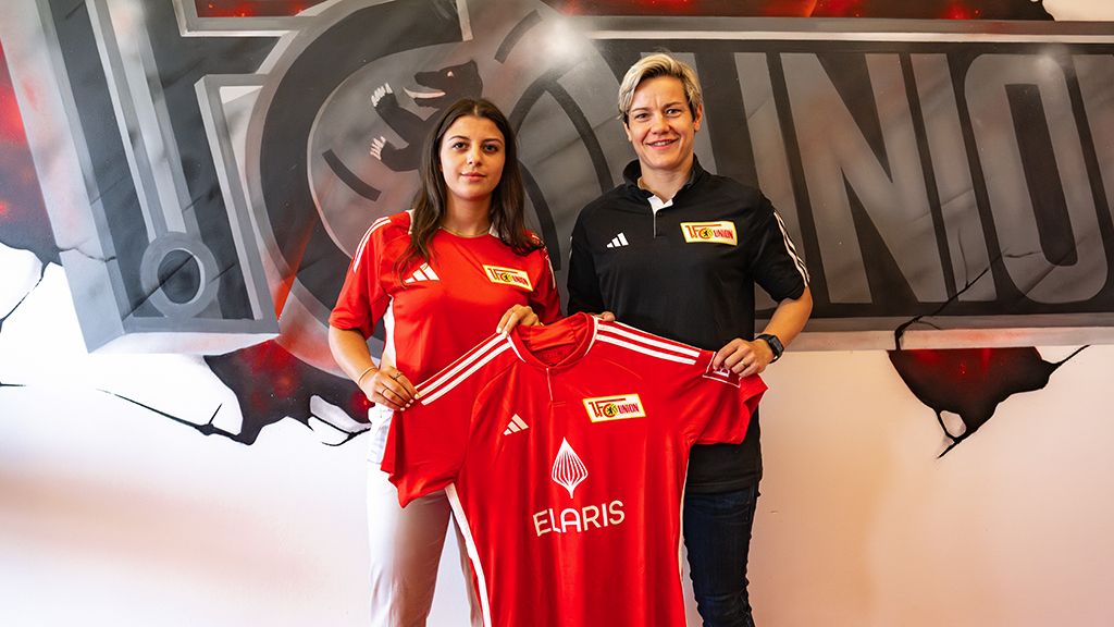 Zwei Frauen halten ein rotes Fußballtrikot mit dem Logo von Union Berlin vor einem Hintergrund mit dem Vereinslogo.
