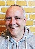 Ein lächelnder Mann mit kurzer, grauer Haarpracht trägt einen grauen Hoodie vor einer gelben Ziegelmauer.