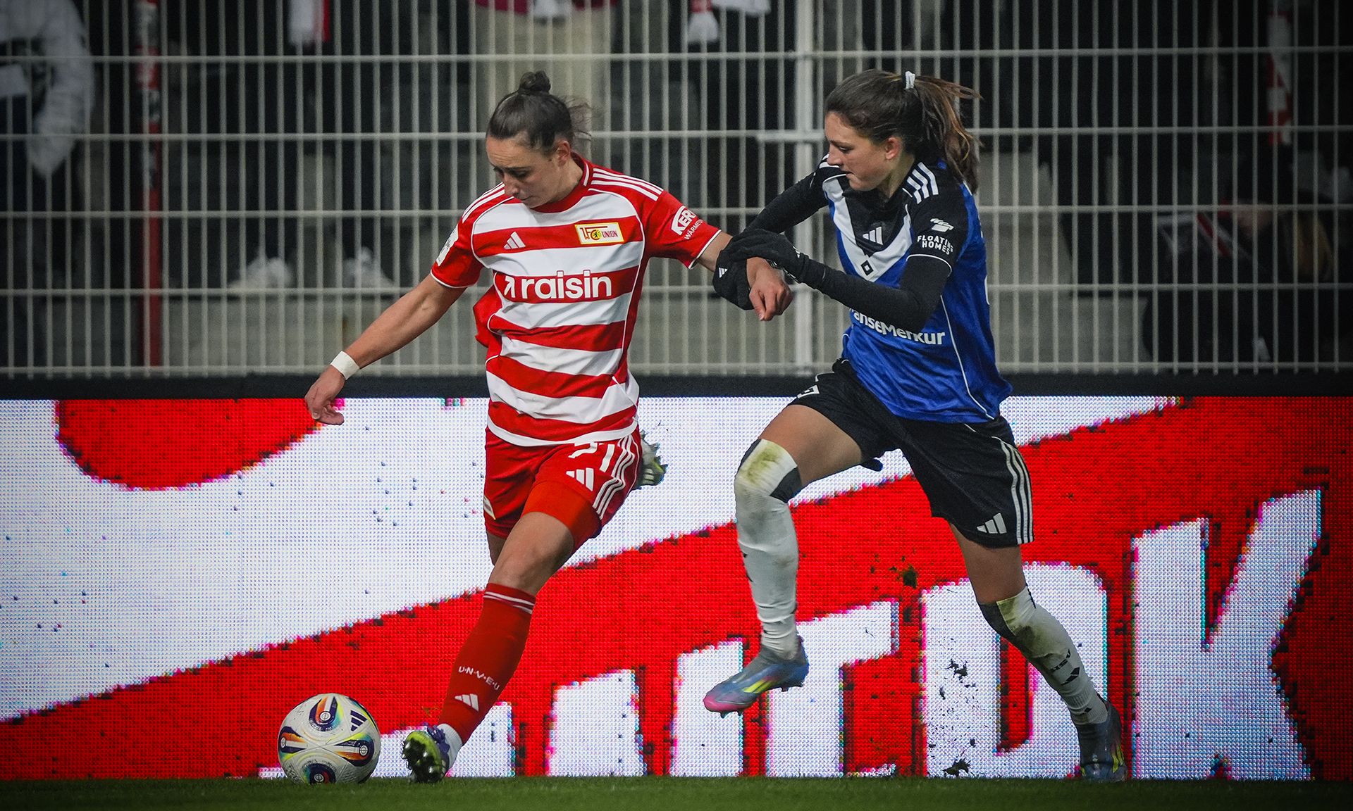 Eine Fußballspielszene mit zwei Spielerinnen, eine in rotem und weißem Trikot, die andere in blau und schwarz, im Wettkampf um den Ball.