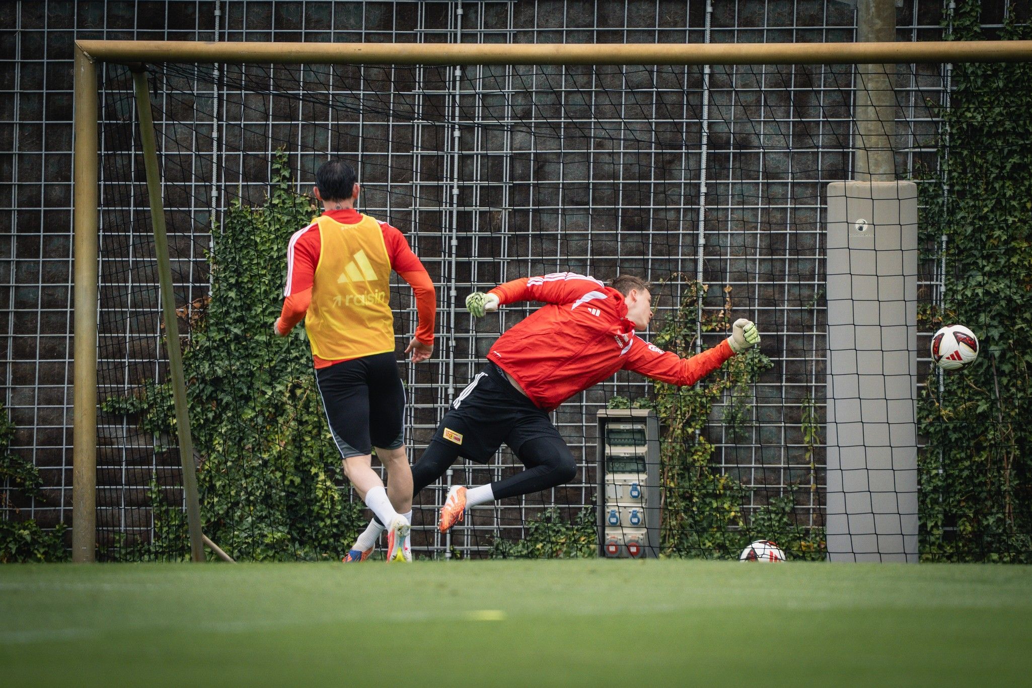 Torwart springt in Richtung Ball, der auf das Tor zufliegt, während ein Spieler im Hintergrund trainiert.