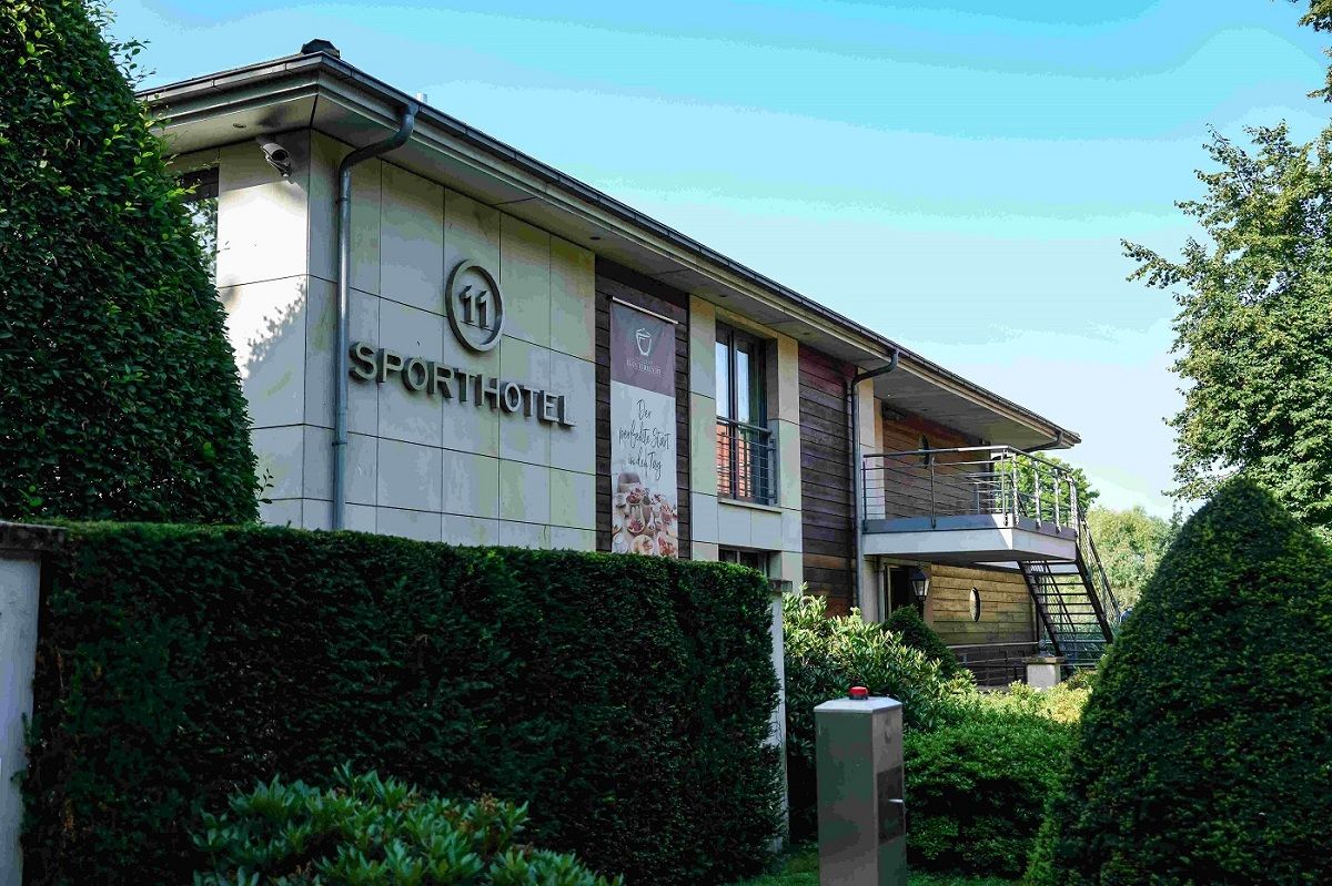 Sporthotel mit moderner Architektur, umgeben von gepflegten Hecken und Bäumen, unter einem klaren blauen Himmel.