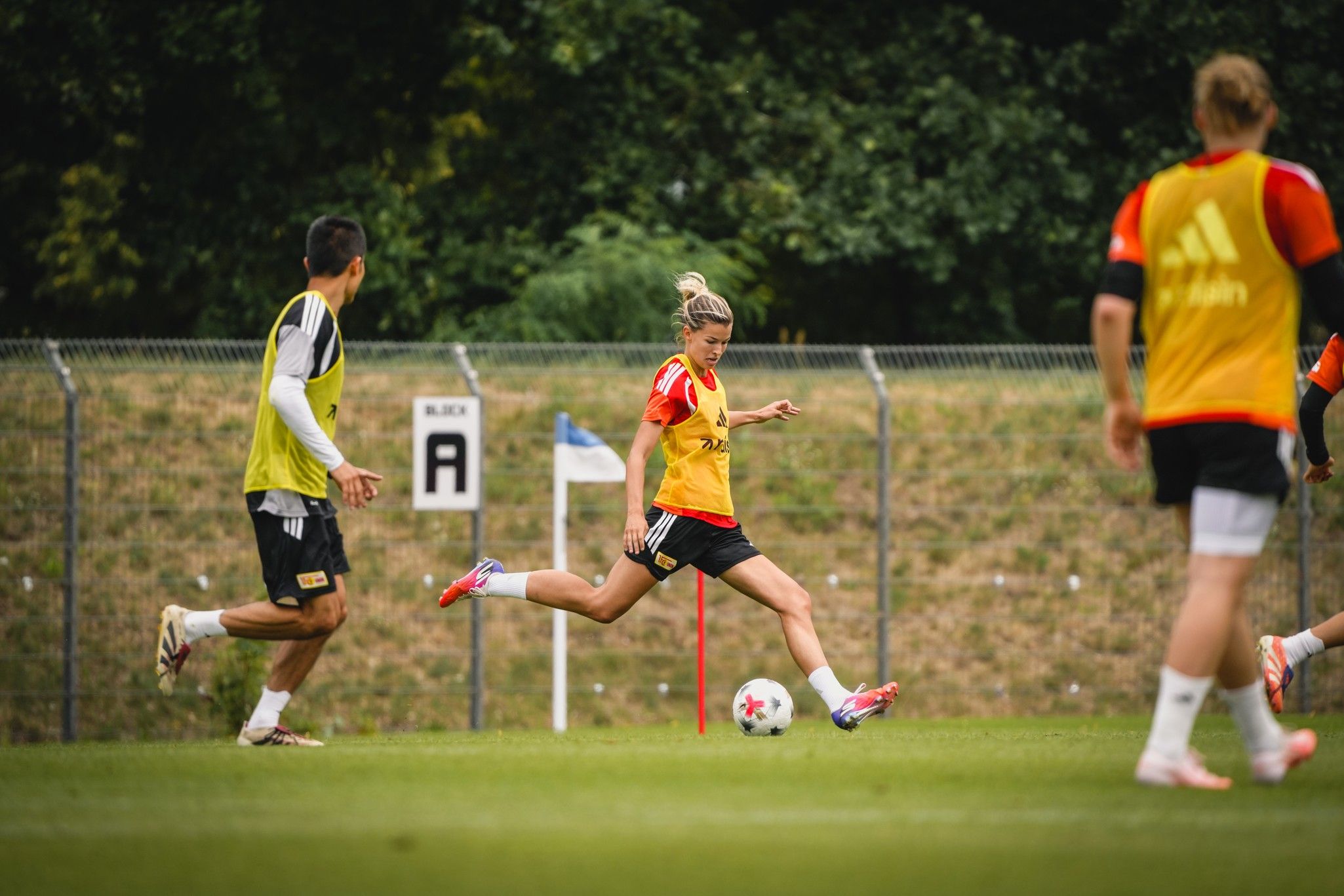 Eine Fußballspielerin in gelber Trainingsweste schießt den Ball, während andere Spieler im Hintergrund trainieren.