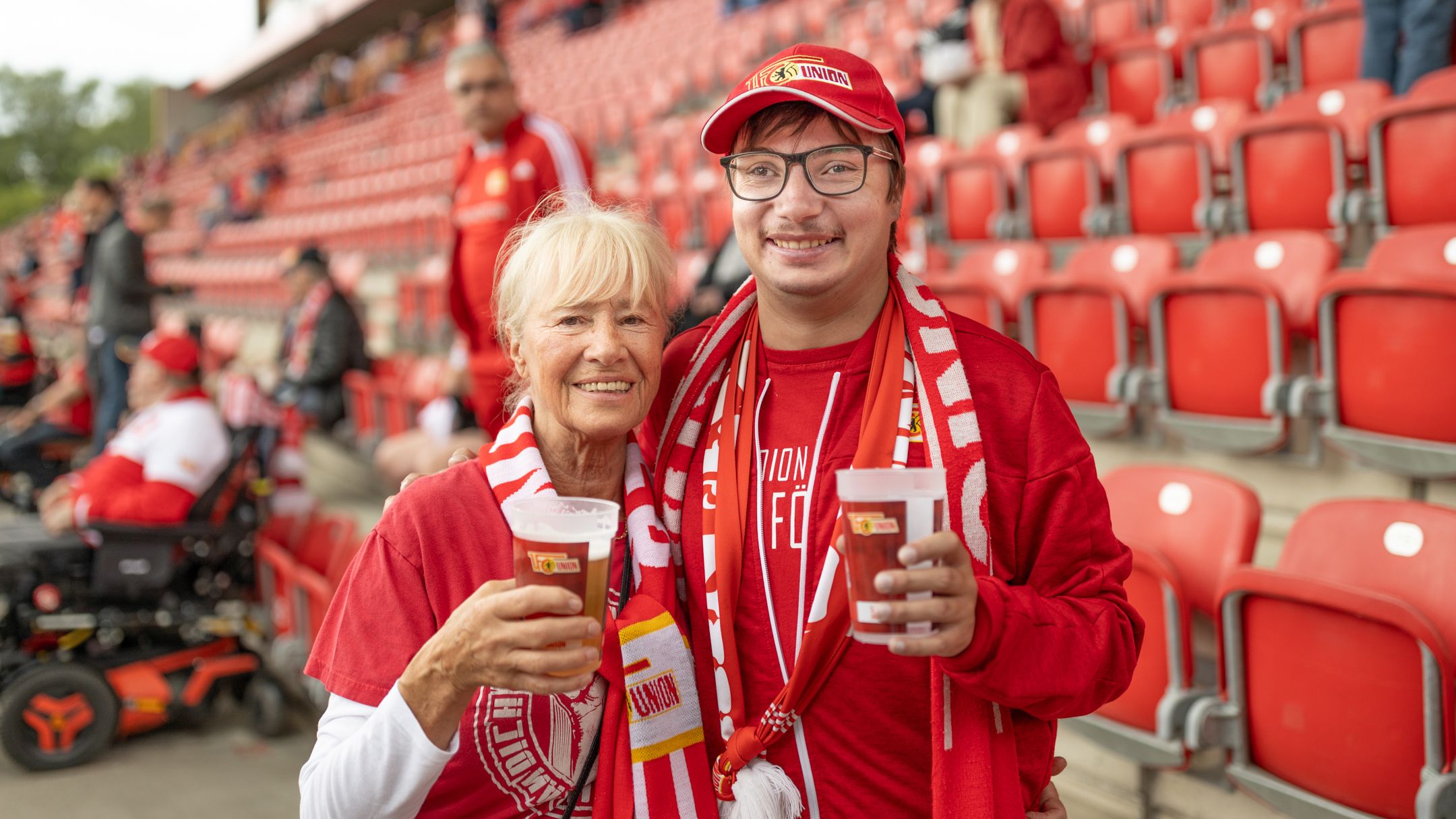 Zwei Fans in roten Outfits halten Getränke und lächeln vor einer leeren Tribüne im Stadion.