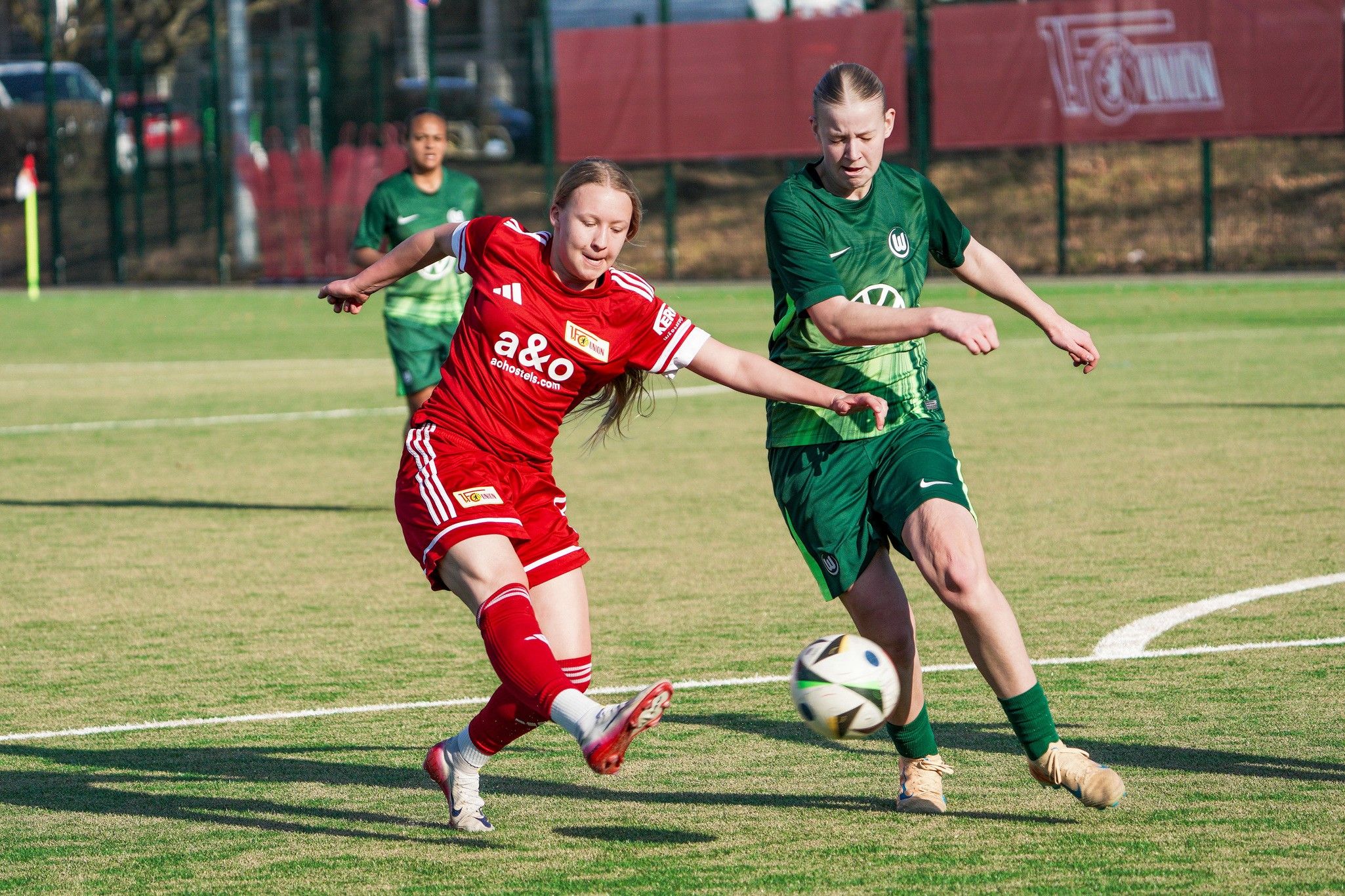 Zwei Fußballspielerinnen im direkten Duell auf einem Rasenplatz, eine in rotem und die andere in grünem Trikot.