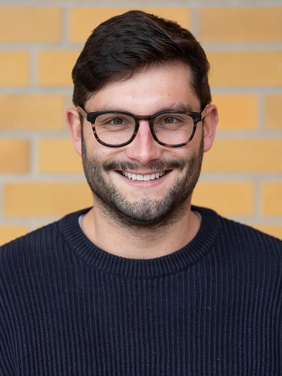 Ein lächelnder Mann mit Brille trägt einen blauen Pullover vor einer gelben Backsteinwand.