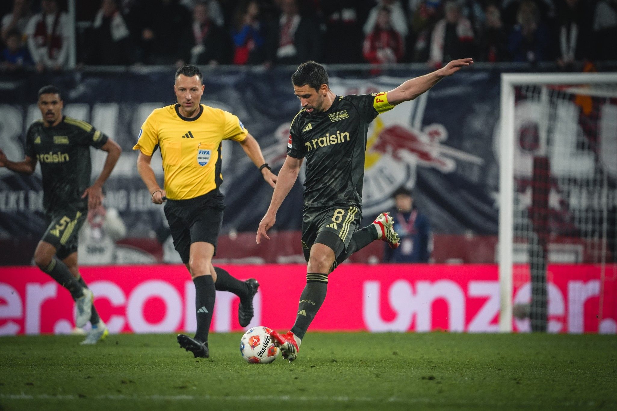 Ein Fußballspieler in schwarz-gelber Kleidung schießt den Ball während eines Spiels. Im Hintergrund ein Schiedsrichter und Fans.