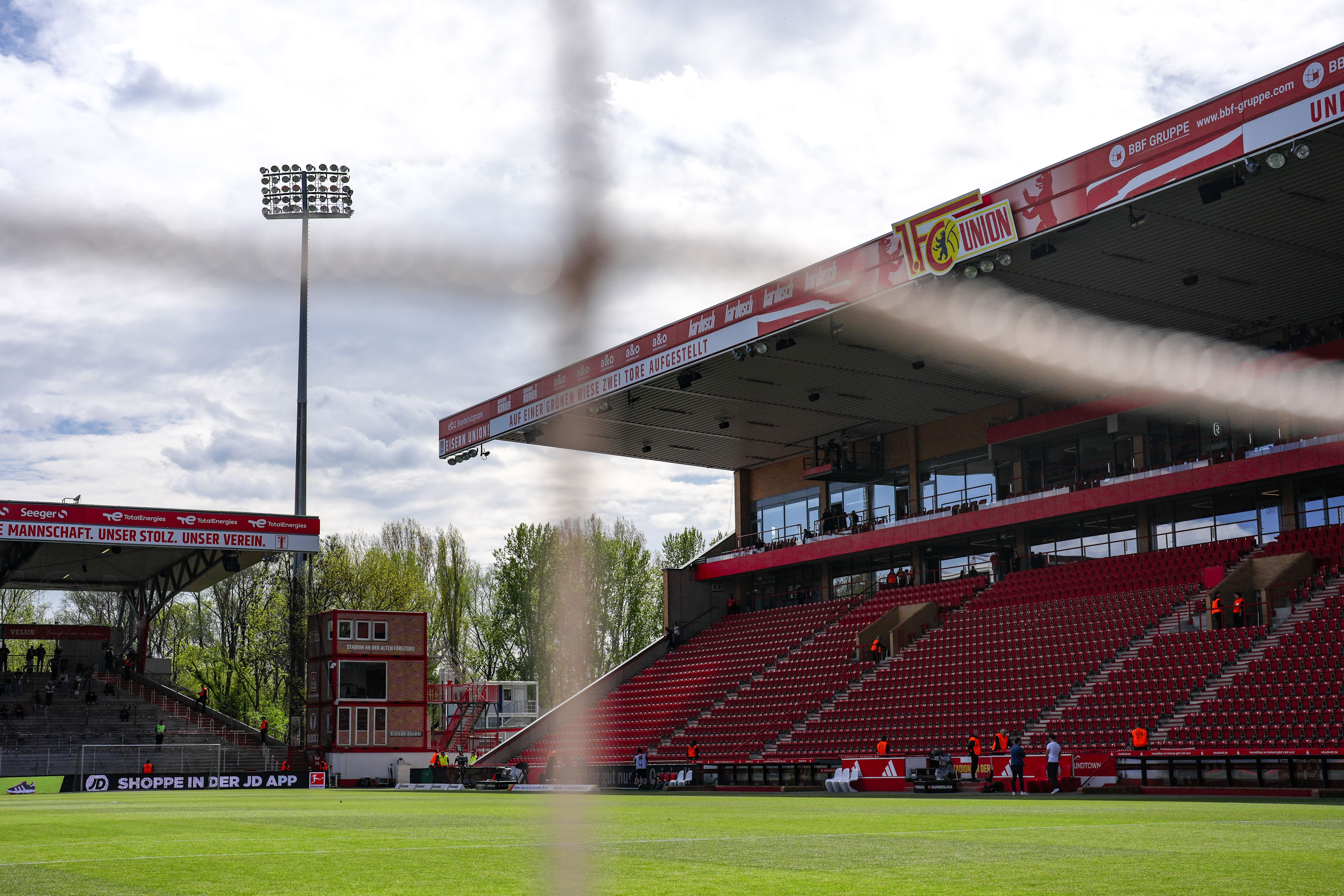 Blick durch ein Fußballtor auf ein leeres Stadion mit roter Bestuhlung und wolkigem Himmel.
