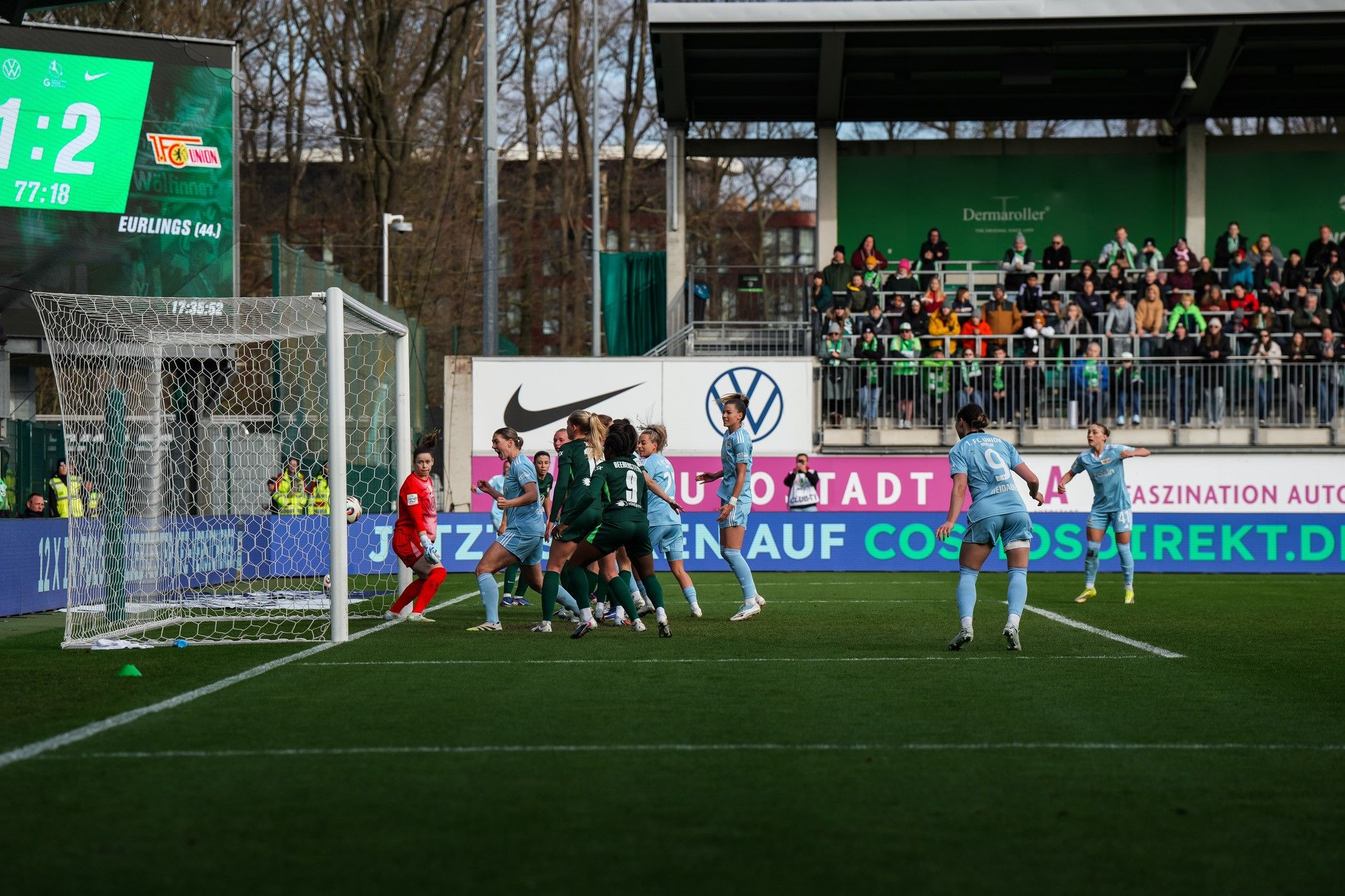 Spielszene im Fußball mit spielenden Mannschaften in grünen und blauen Trikots, Torwart und Torrahmen im Fokus.