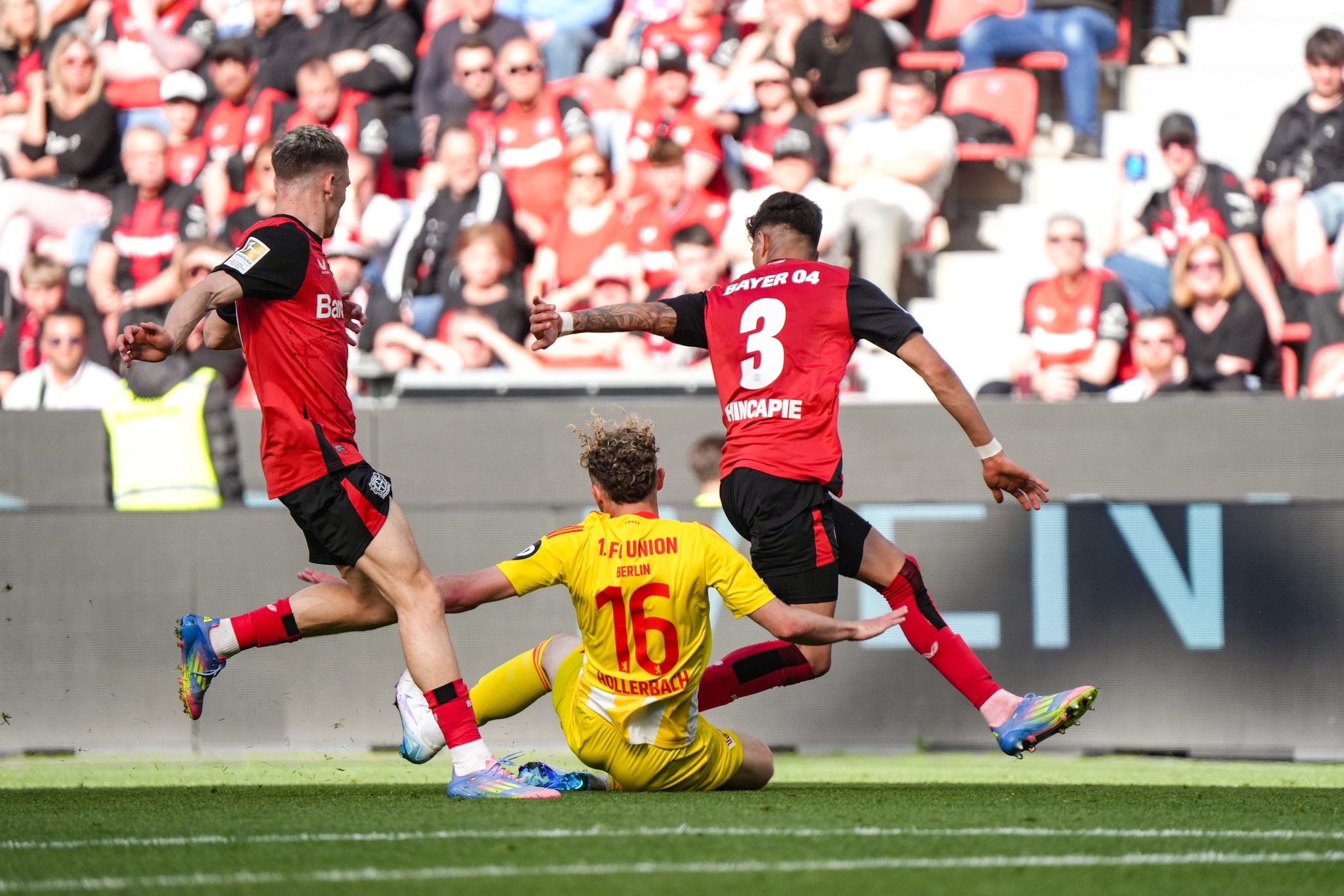 Zwei Fußballspieler in roten Trikots stehen vor einem am Boden liegenden Spieler in gelbem Trikot auf einem Stadionrasen.