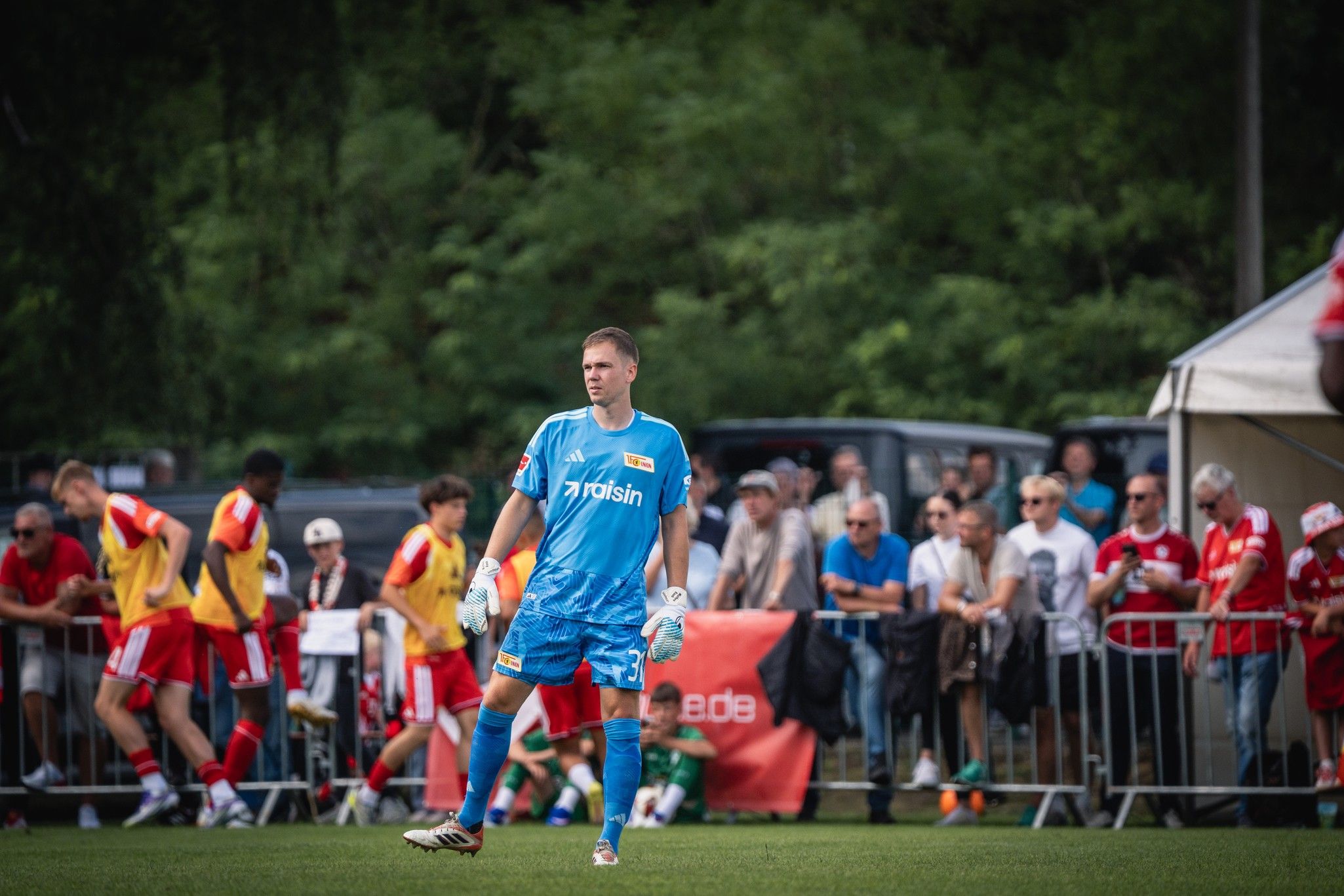 Ein Fußballtorwart in blauer Ausrüstung steht auf dem Spielfeld, während Zuschauer und Spieler an der Seitenlinie zuschauen.